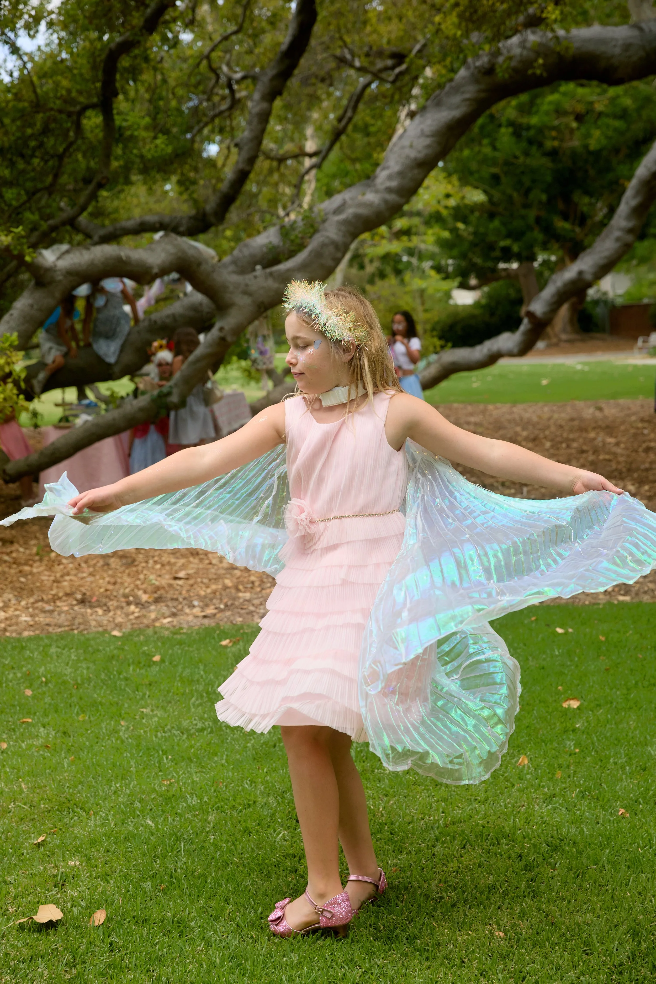 A joyful young girl in a pink dress and sheer blue fairy wings running happily through a grassy park.