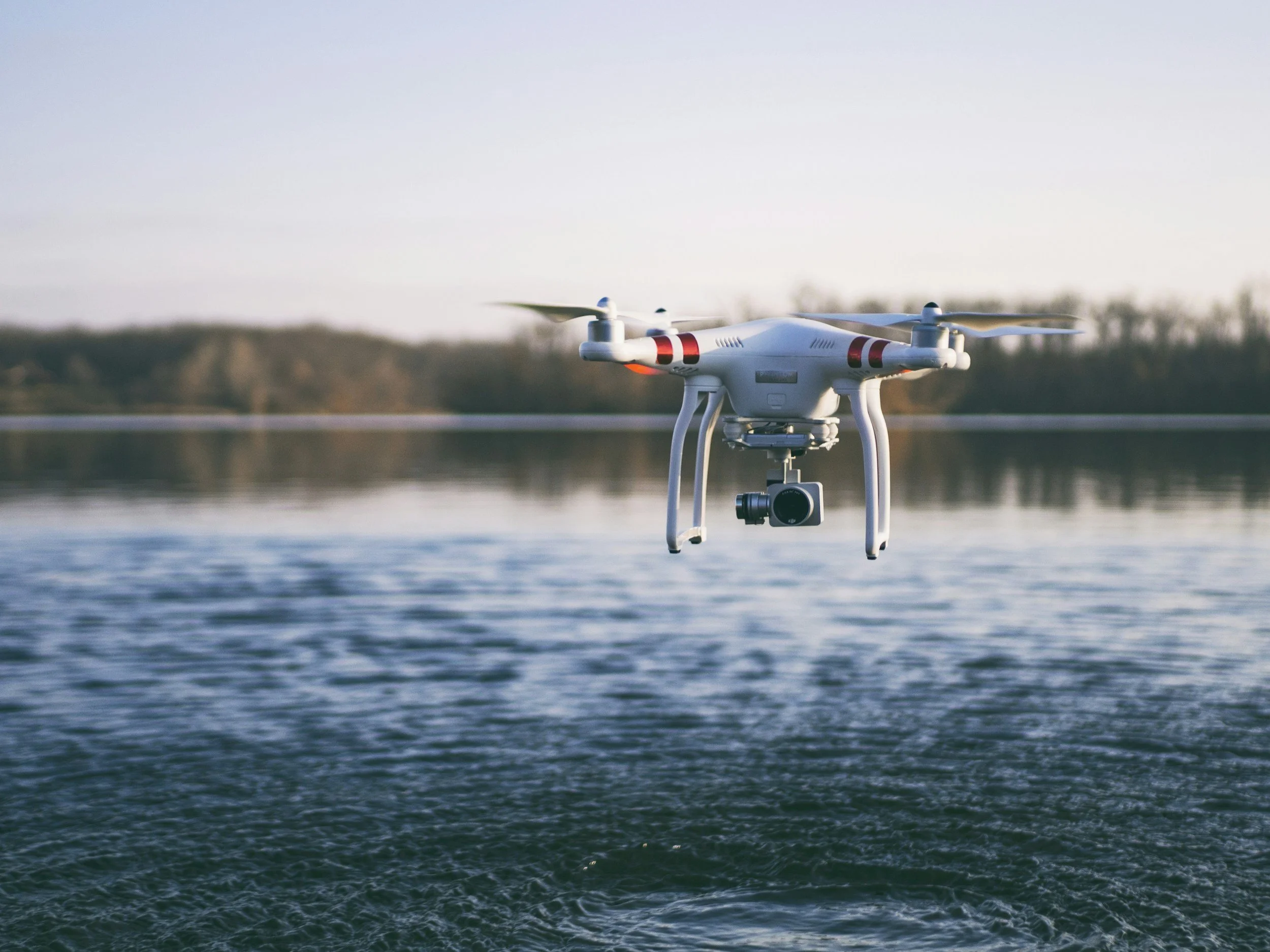 A white quadcopter drone flying over a body of water during sunset or sunrise, with a blurred background of trees and hills.