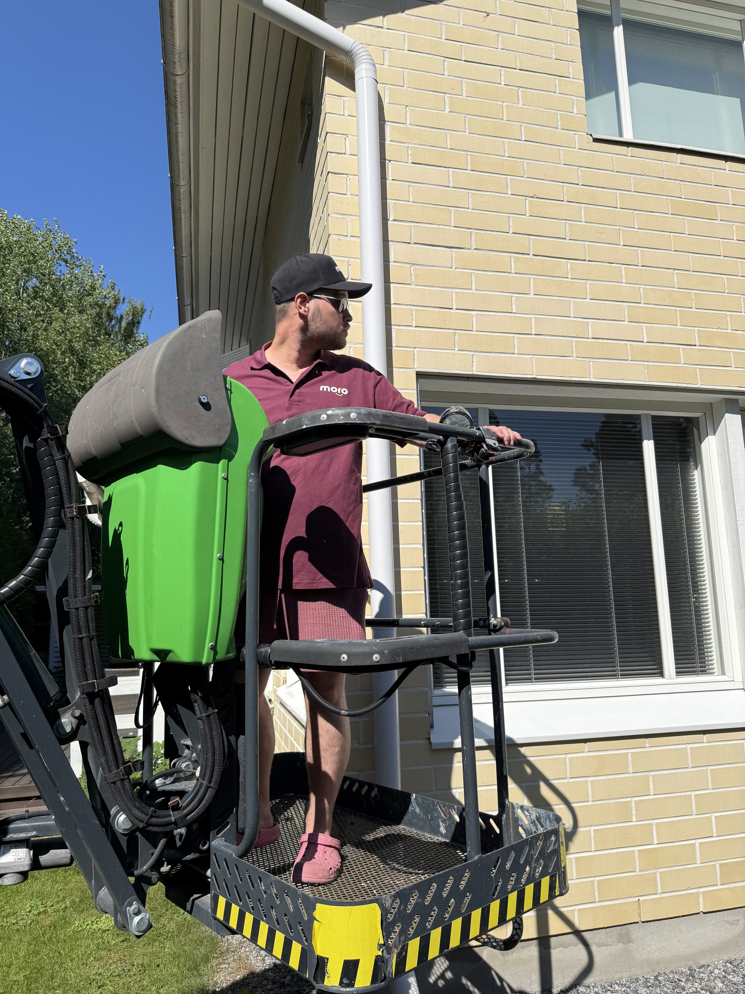 Mies työnteossa rakennuksen vieressä, joka on rakennettu keltaisesta tiilestä. Hän käyttää tummia aurinkolaseja, mustaa lippalakkia ja viininpunaista t-paitaa.