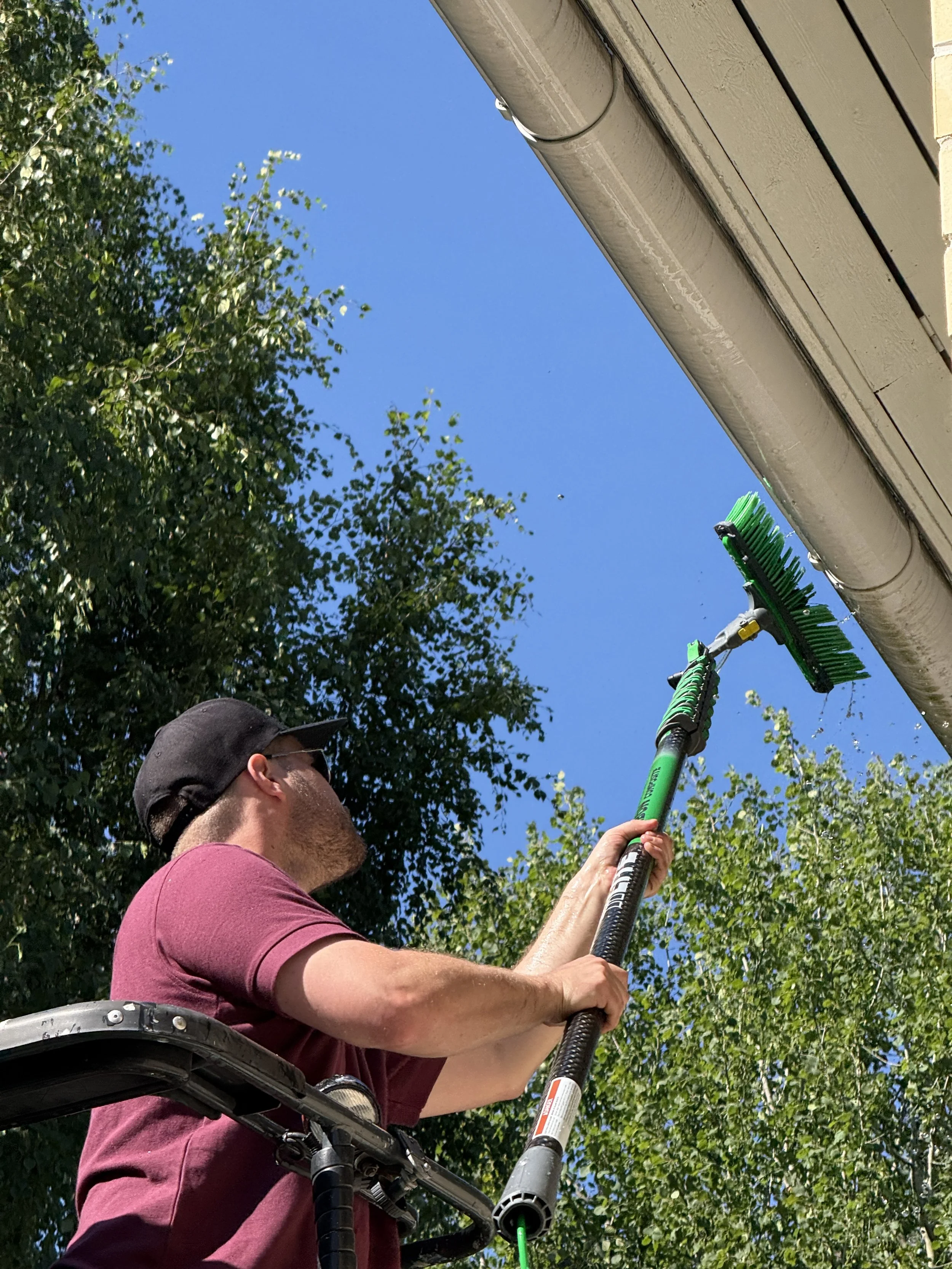 Henkilö käyttää varrellista pesuharjaa henkilönostimen kyydissä, puhdistaakseen rännejä. Taustalla on vihreitä puita ja kirkas sininen taivas.