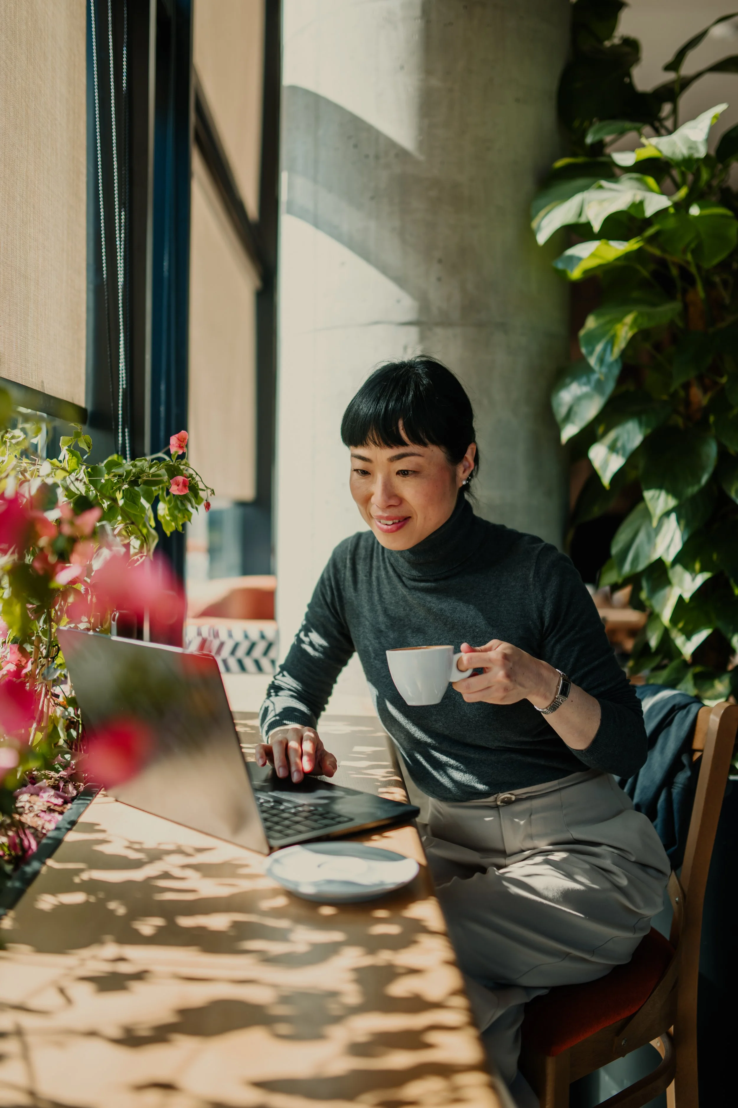 A woman with short black hair, wearing a gray turtleneck top and light-colored pants, is sitting at a wooden table in a cafe. She is holding a white coffee cup in her right hand and using a laptop with her left hand. The cafe has large windows, green plants, and pink flowers on the table, with sunlight casting shadows on the table.