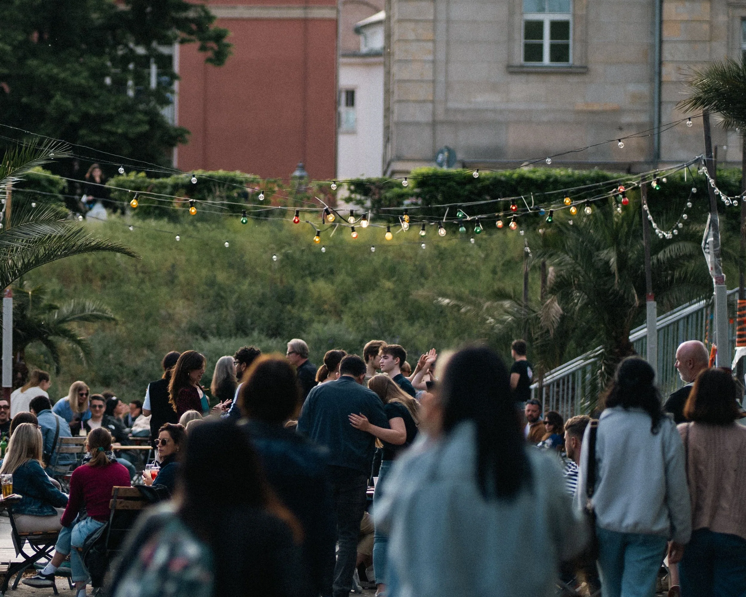 A lively outdoor gathering of diverse people socializing, with string lights hanging above, greenery, and buildings in the background.