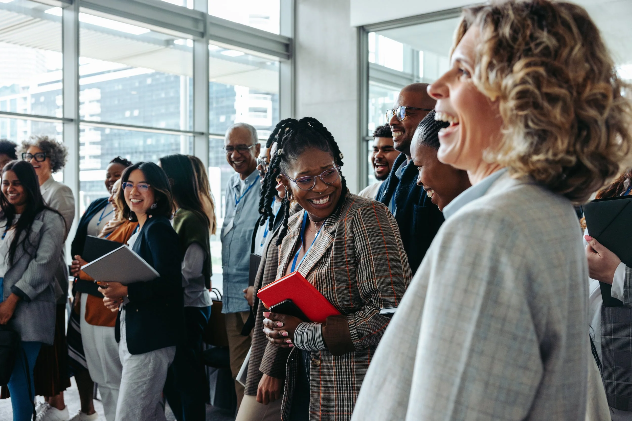 Group of diverse people smiling and laughing at a conference or seminar in a modern building with large windows.