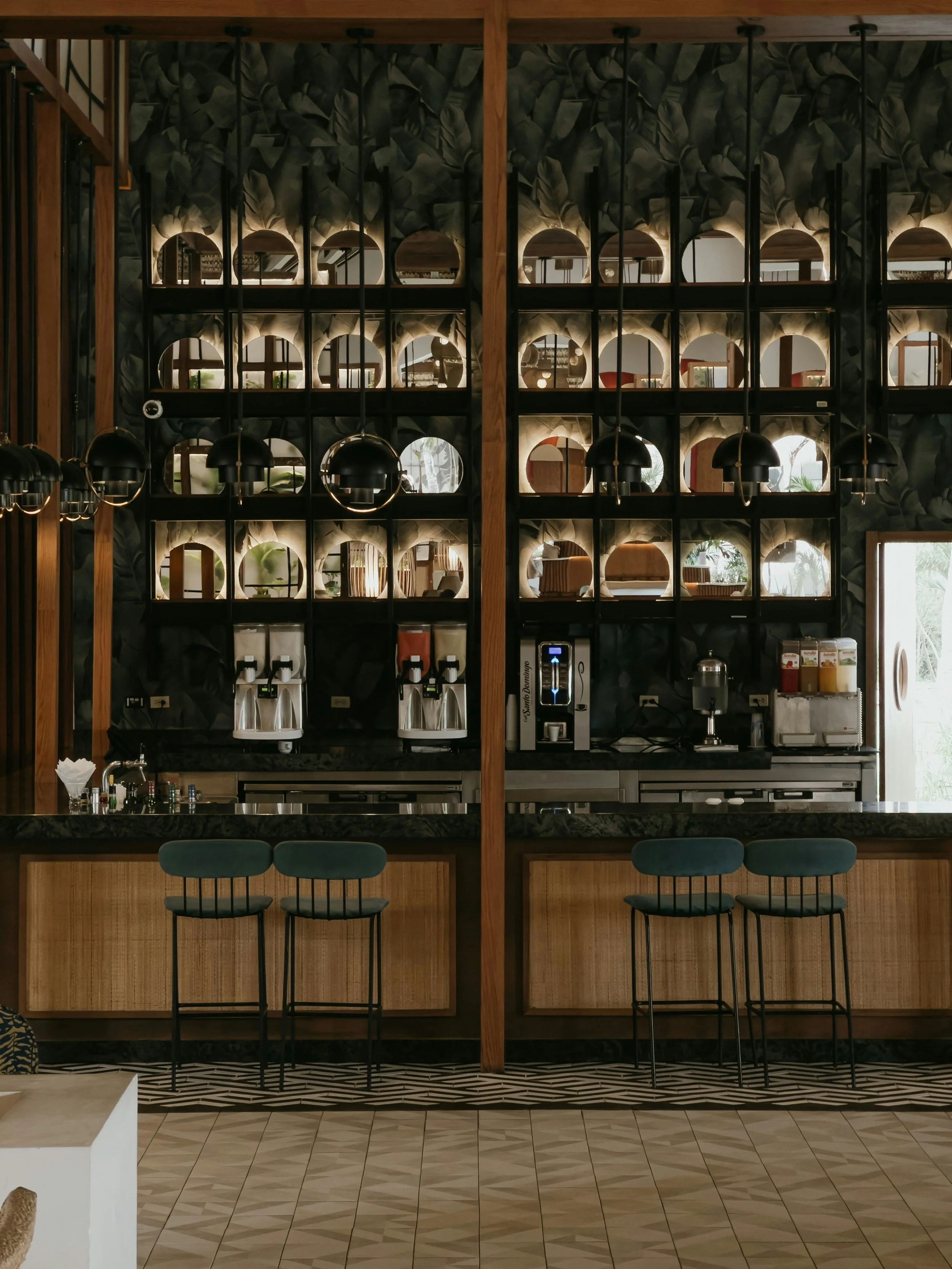 Interior view of a modern cafe or restaurant with a bar, decorated with a black and green leafy wallpaper pattern, featuring a large mirror wall, hanging black pendant lights, and teal barstools with black frames.