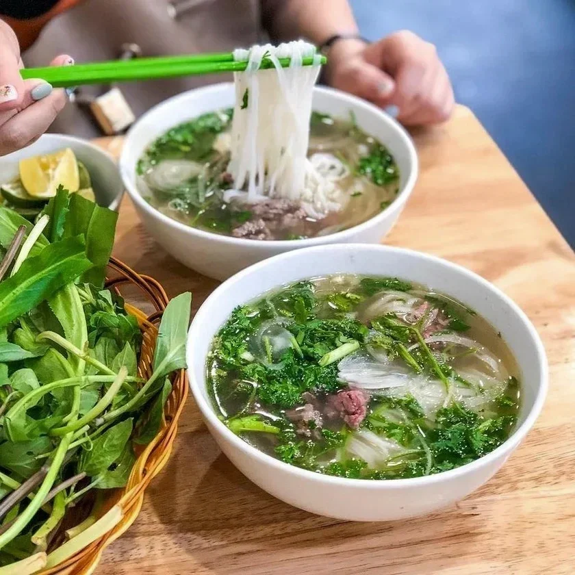 Two bowls of Vietnamese pho with beef, green herbs, and rice noodles on a wooden table, with a basket of fresh herbs and lemon wedges nearby.