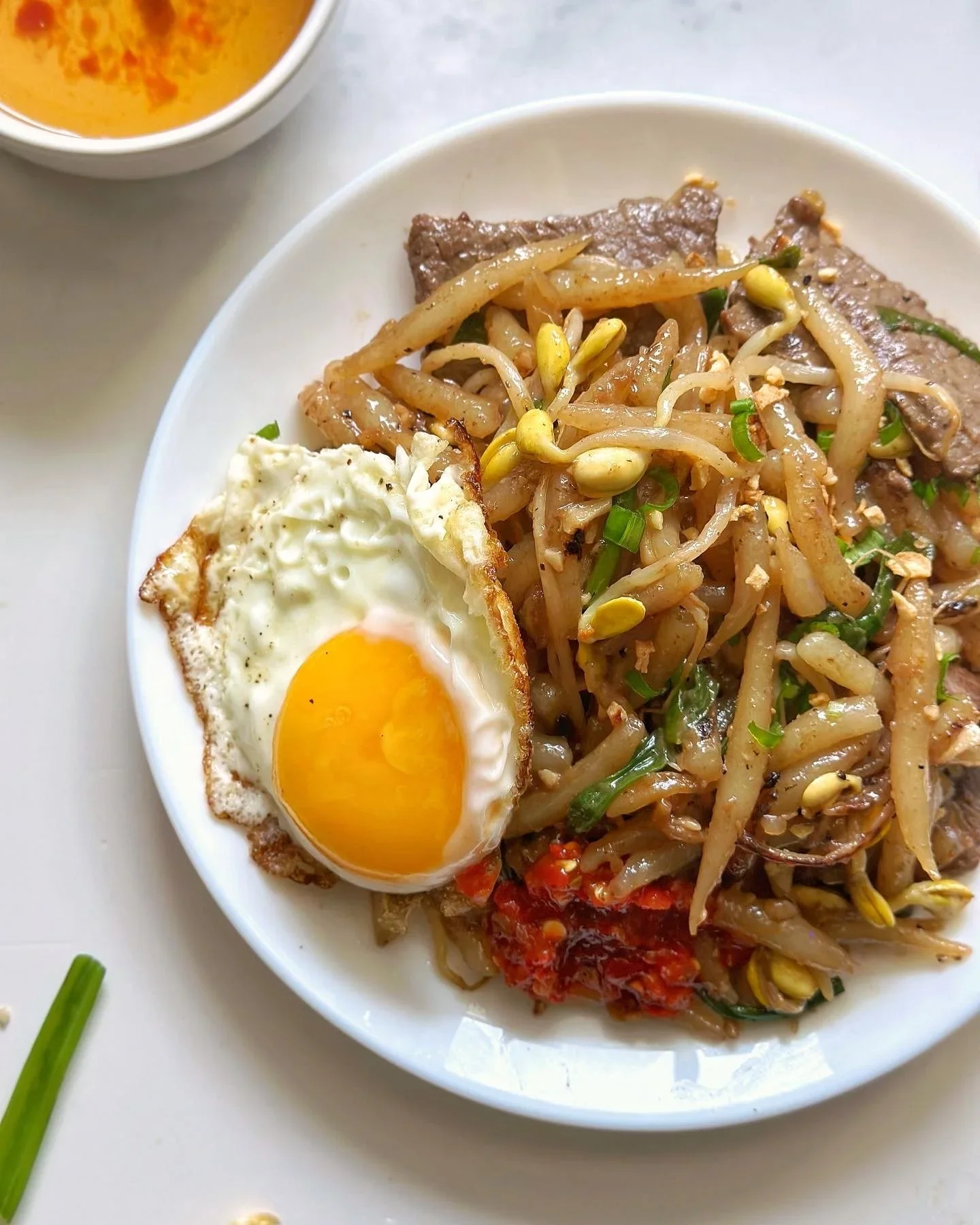 Close-up of a white bowl containing Asian stir-fried bean sprouts, beef slices, a fried egg with a runny yolk, and red chili sauce. A small bowl of orange-colored soup is partially visible at the top left corner.