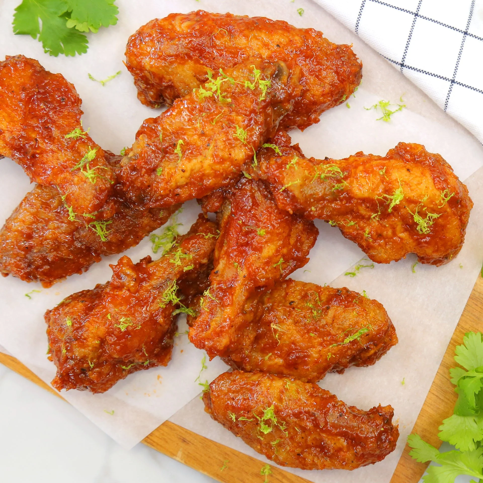 Close-up of crispy, saucy chicken drumsticks garnished with lime zest, placed on parchment paper on a wooden board, with cilantro and a checkered napkin nearby.