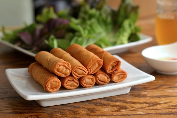 A plate of six crispy egg rolls on a white rectangular dish, with a side of salad greens in the background and a glass of beer to the right.