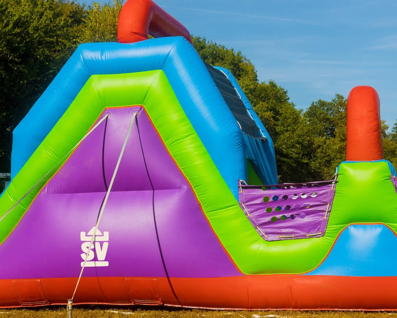 Colorful inflatable bounce house or set up outdoors on grass, with trees and a blue sky in the background.