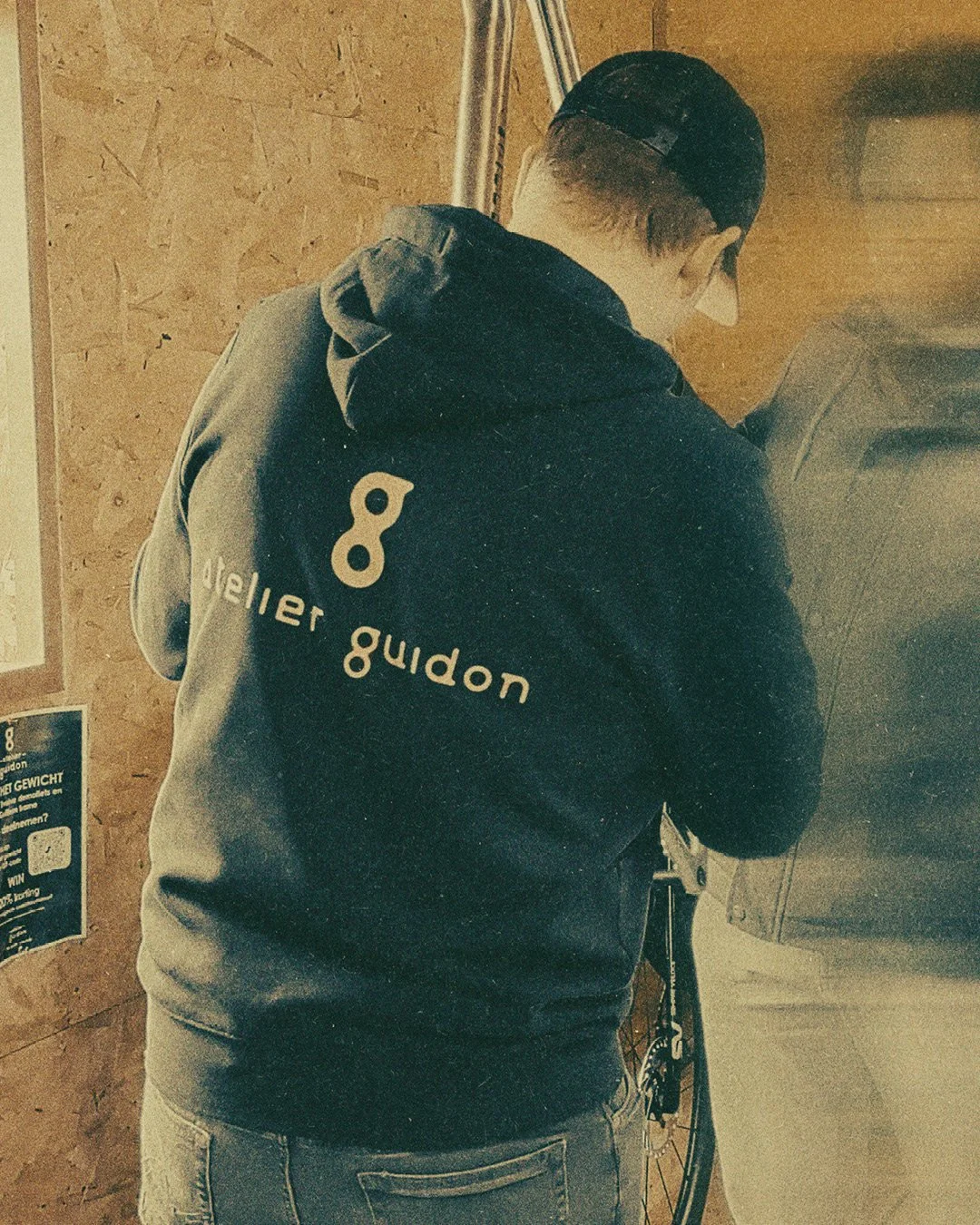 Back of a person wearing a black hoodie with a white number 8 and the words 'aller guidon' written on it, leaning forward and working on a bicycle in a workshop with wooden walls.