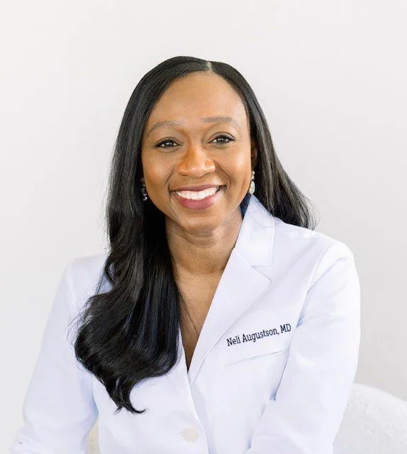A woman smiling, wearing a white lab coat with a name tag that reads Neli Augustson, MD, and earrings, standing against a plain white background.
