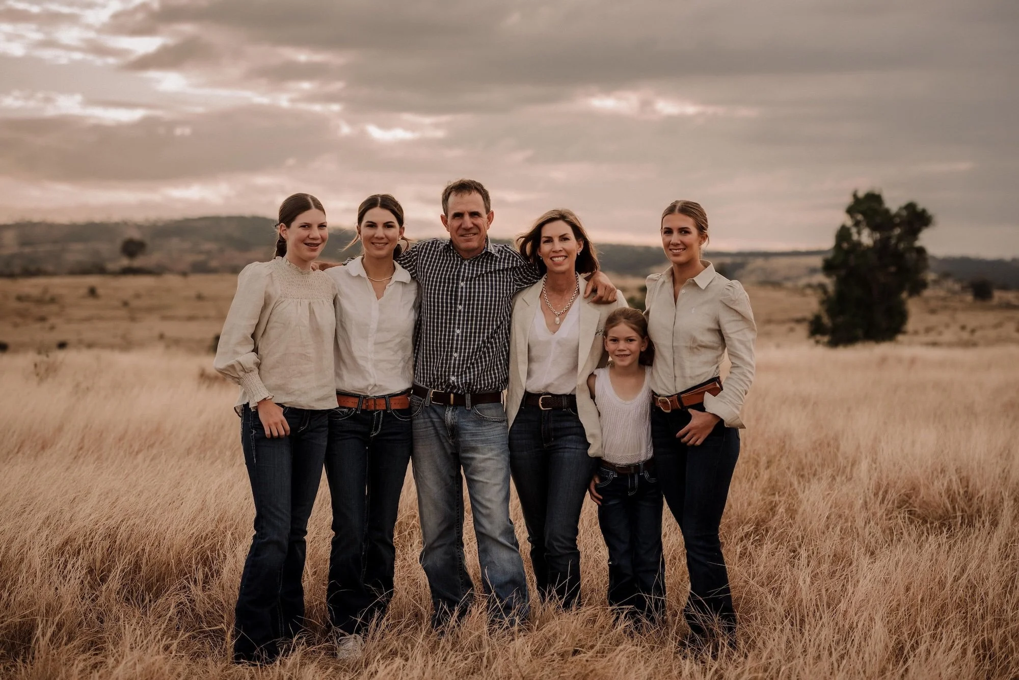 Family of six, including two young girls, posing together outdoors in a field with tall grass under a cloudy sky.
