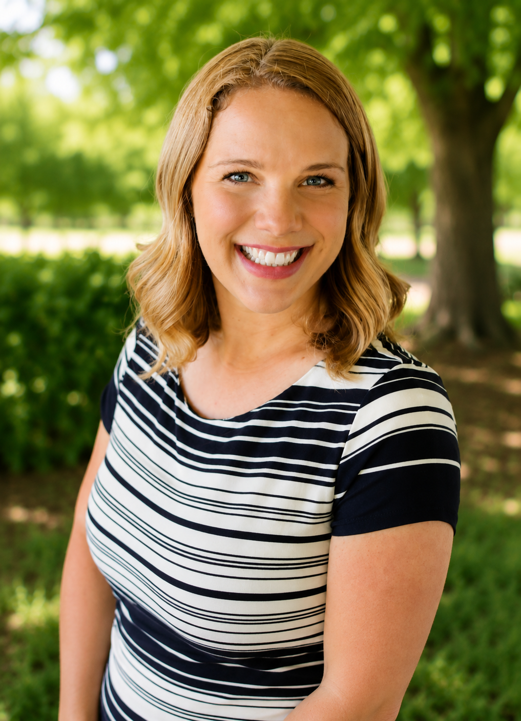 A woman with blonde, wavy hair smiling outdoors with green trees and bushes in the background.