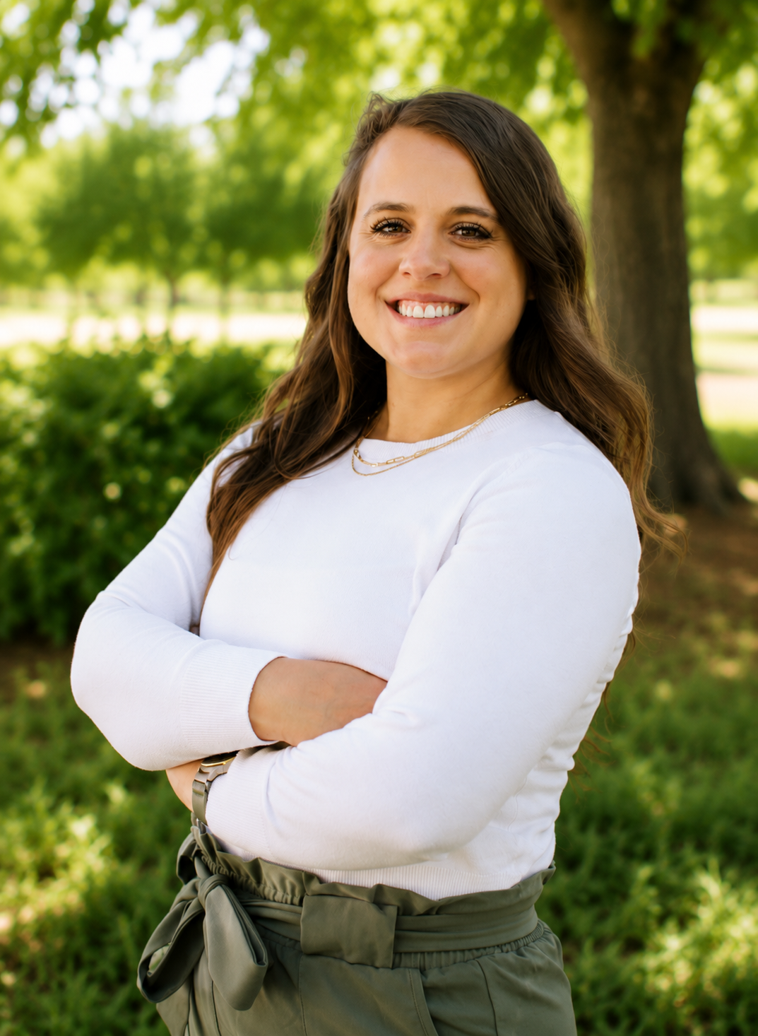 A woman with long, wavy brown hair, smiling, wearing a white long-sleeve shirt, layered necklaces, and green pants with a tied waist, standing outdoors in front of green trees and bushes on a sunny day.