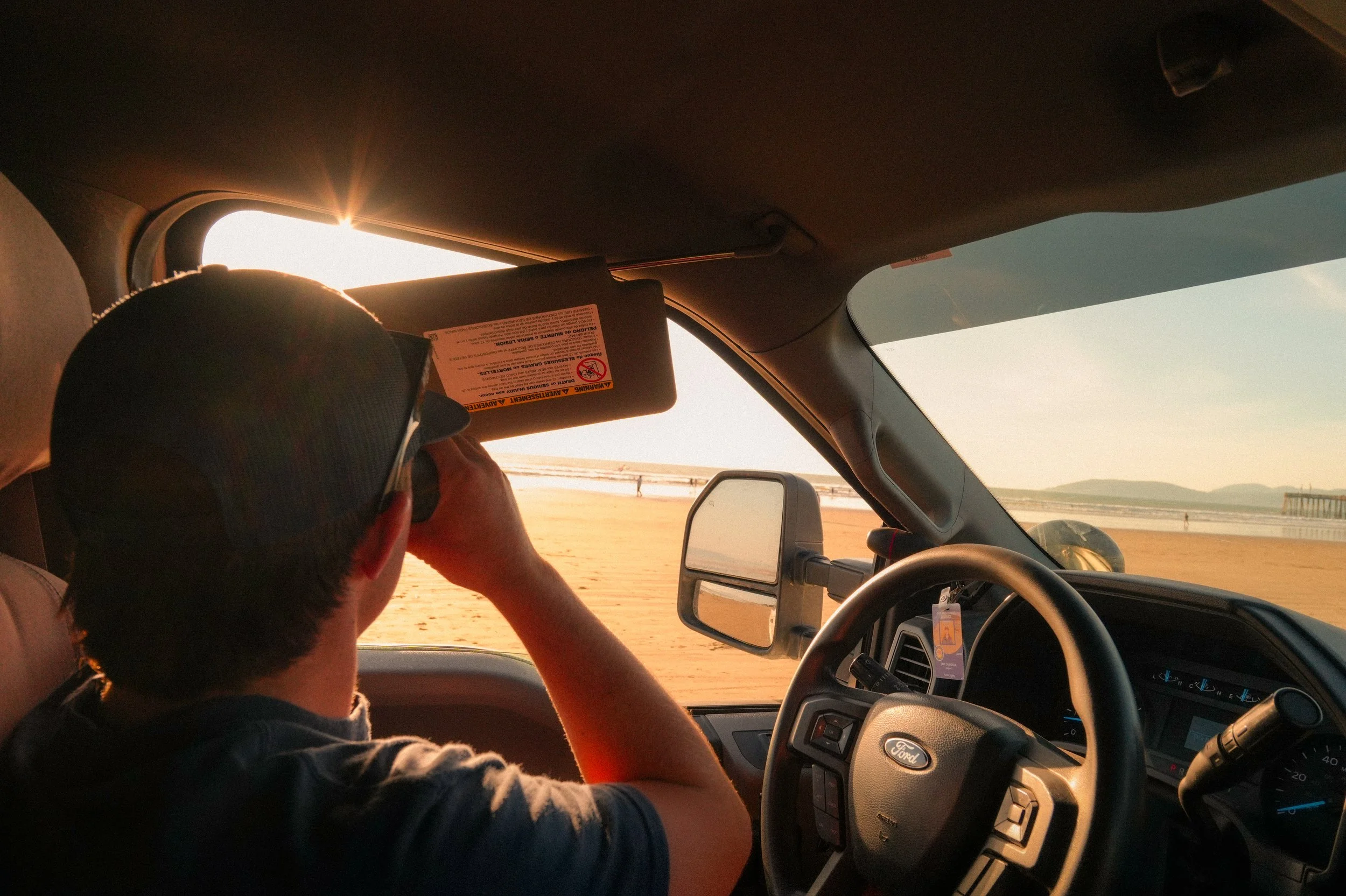 Inside a vehicle, a person wearing a cap is holding a phone or camera, taking a picture of the sunset through the windshield. The sunset is casting a warm glow, with an ocean and a pier visible outside.