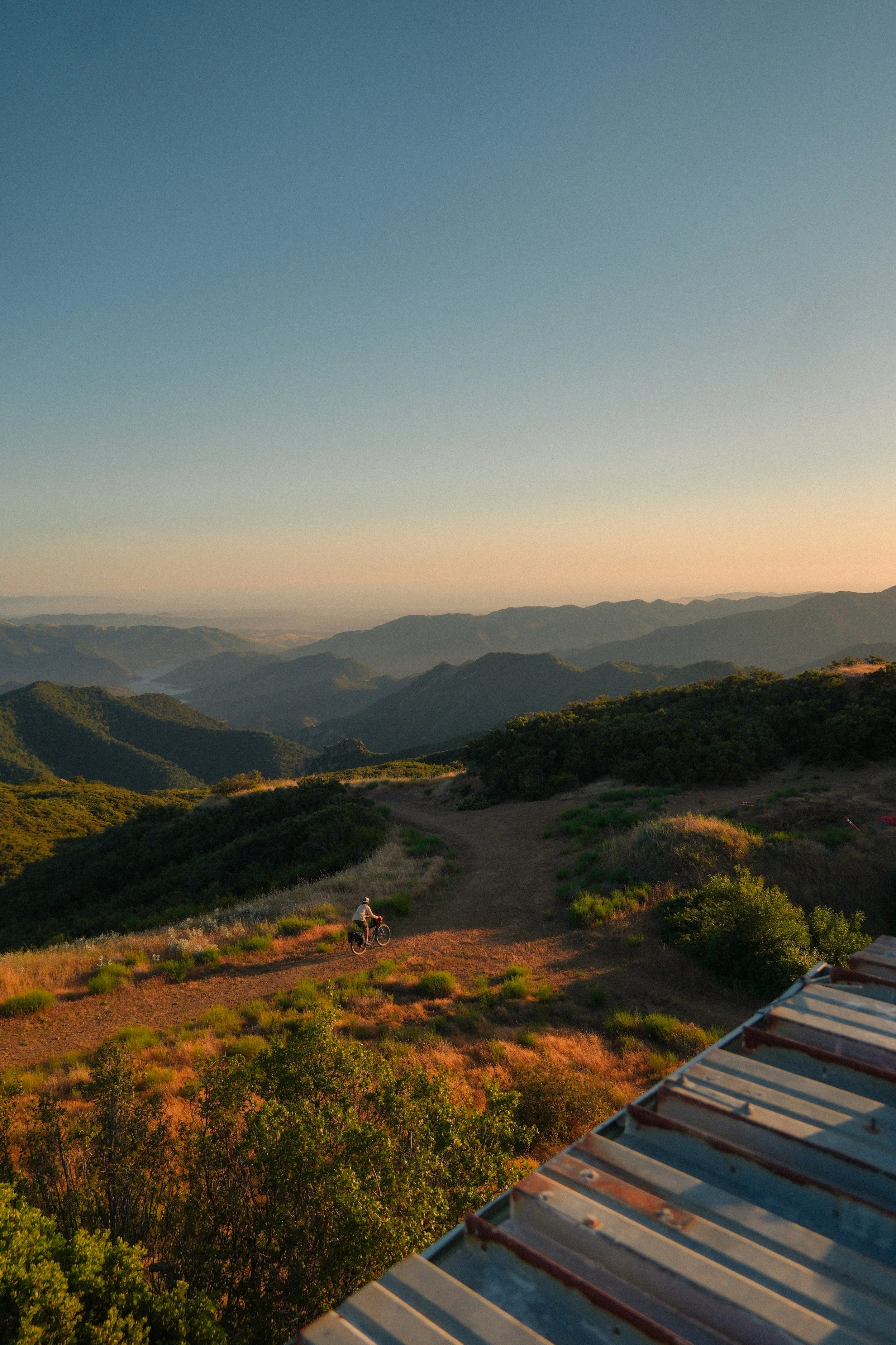 A person riding a bicycle up a dirt trail through a hilly landscape with green shrubs and trees during sunset or sunrise, with mountains in the distance and a partly cloudy sky.