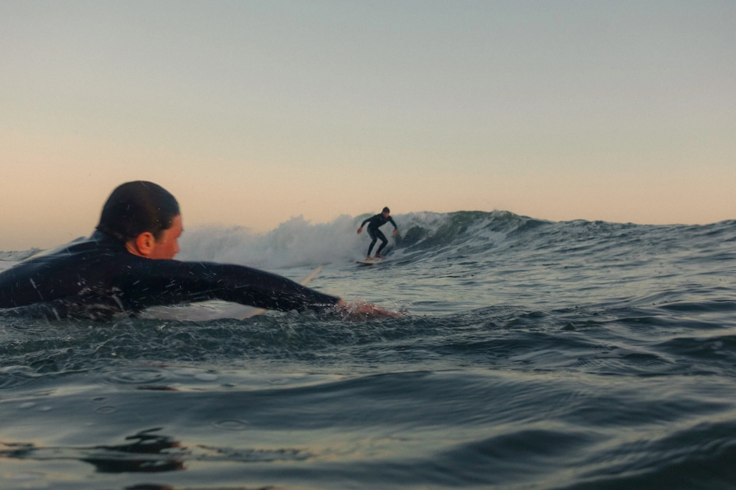 A person is lying on a surfboard in the water, close to the camera, with another person riding a wave in the distance. The scene is set during sunset or sunrise, with a calm sky and distant clouds.