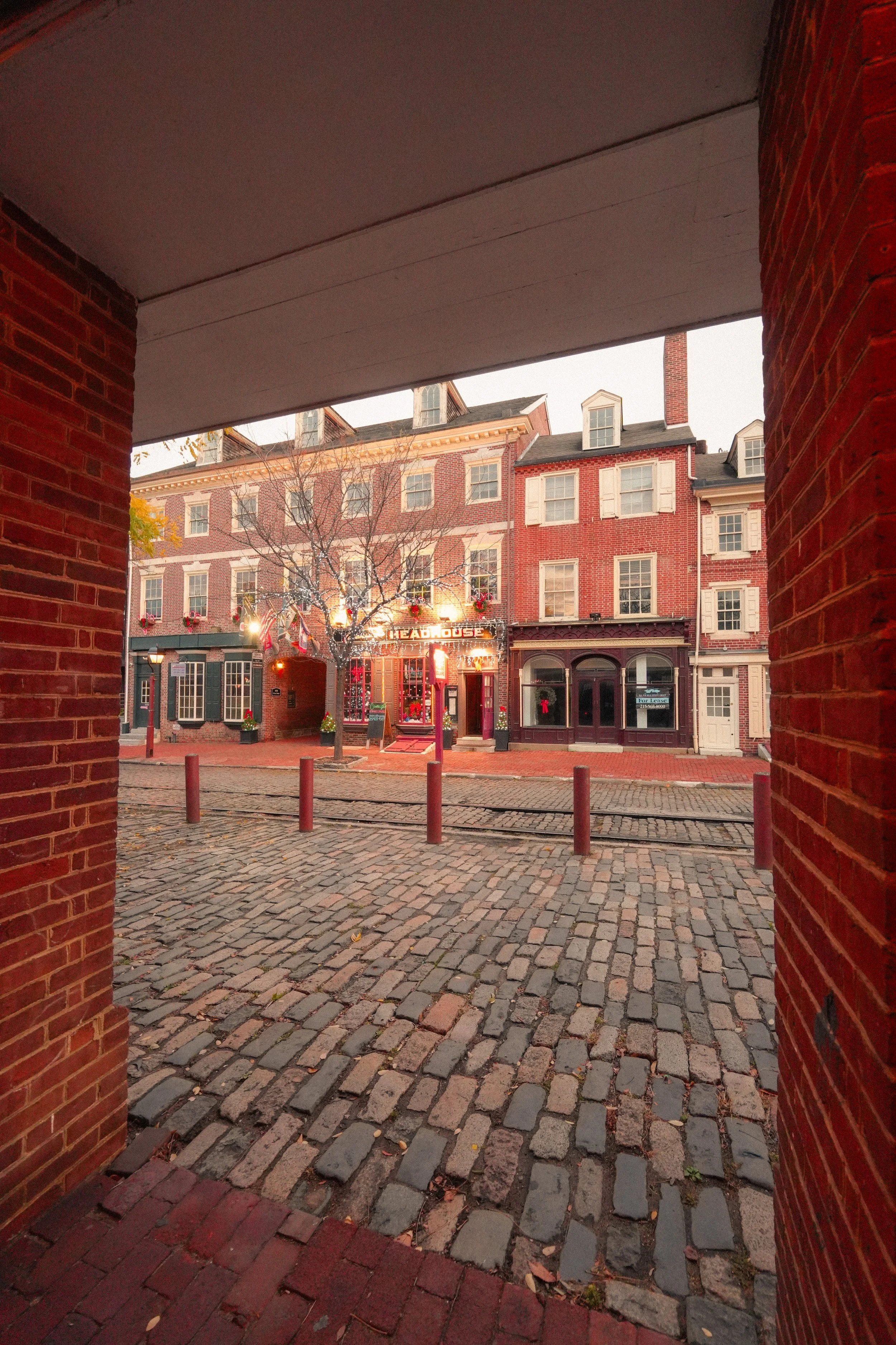 View of a city street through a brick archway, featuring cobblestone pavement, a leafless tree, and buildings with windows and storefronts, some decorated for the holiday season.