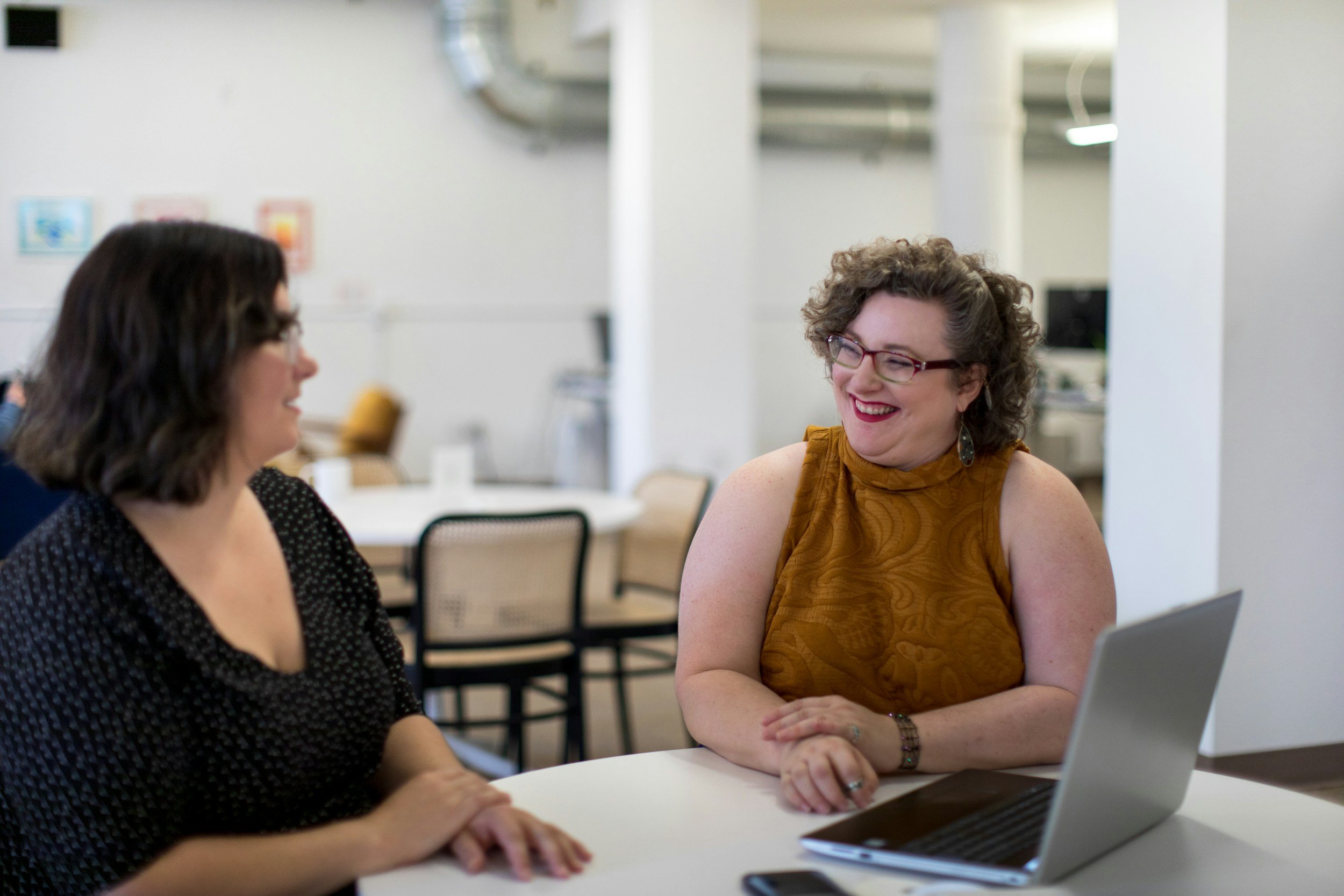 Two women are sitting at a table and engaging in a conversation, smiling. One woman has curly hair, glasses, and is wearing a sleeveless orange top. The other woman has dark hair and is wearing a black polka dot top. There are chairs and tables in the background of a bright, modern office or conference space.