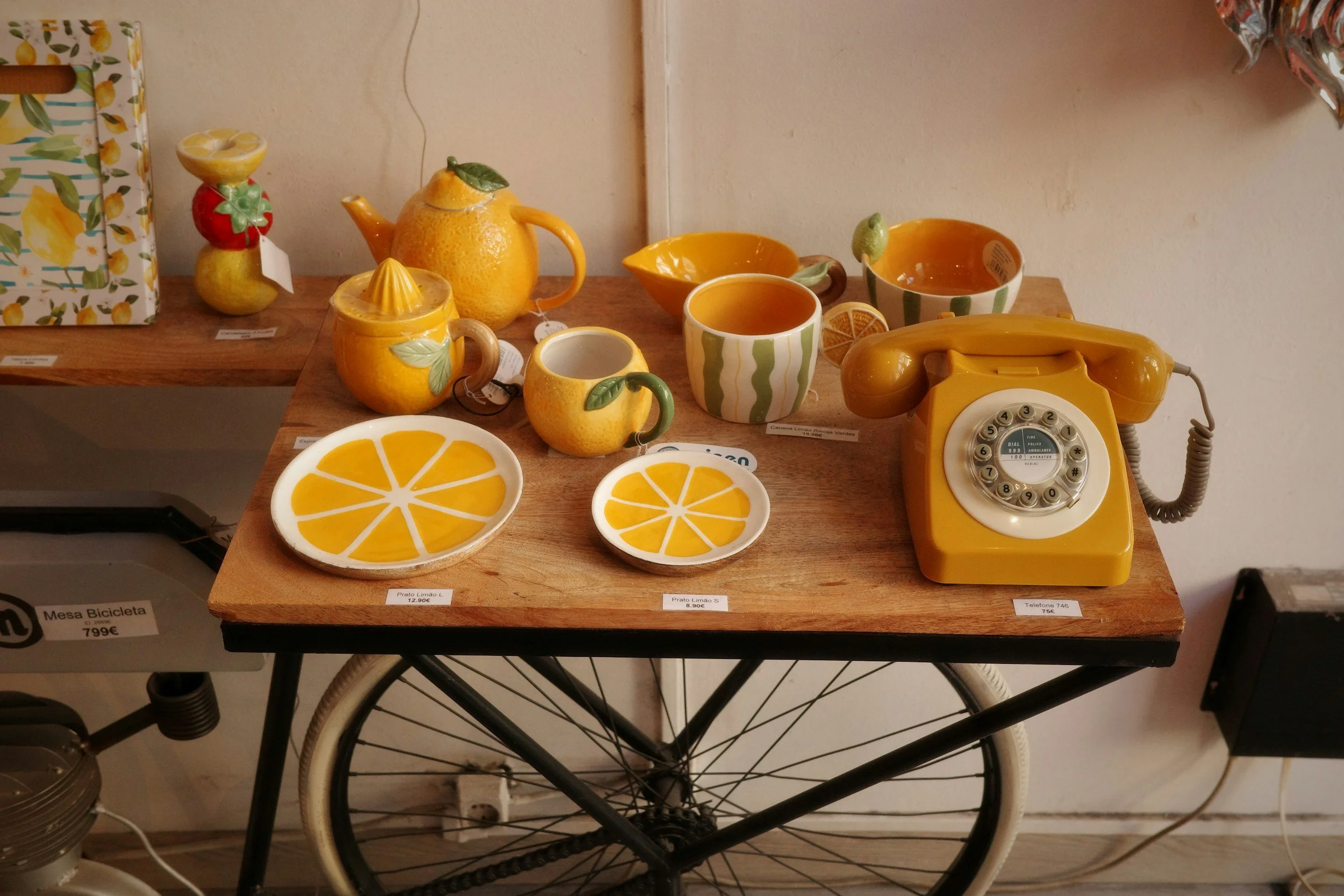 Display of yellow ceramic kitchenware with orange patterns, including cups, bowls, a teapot with a lemon-shaped lid, a citrus juicer, and a rotary phone, arranged on a wooden table.