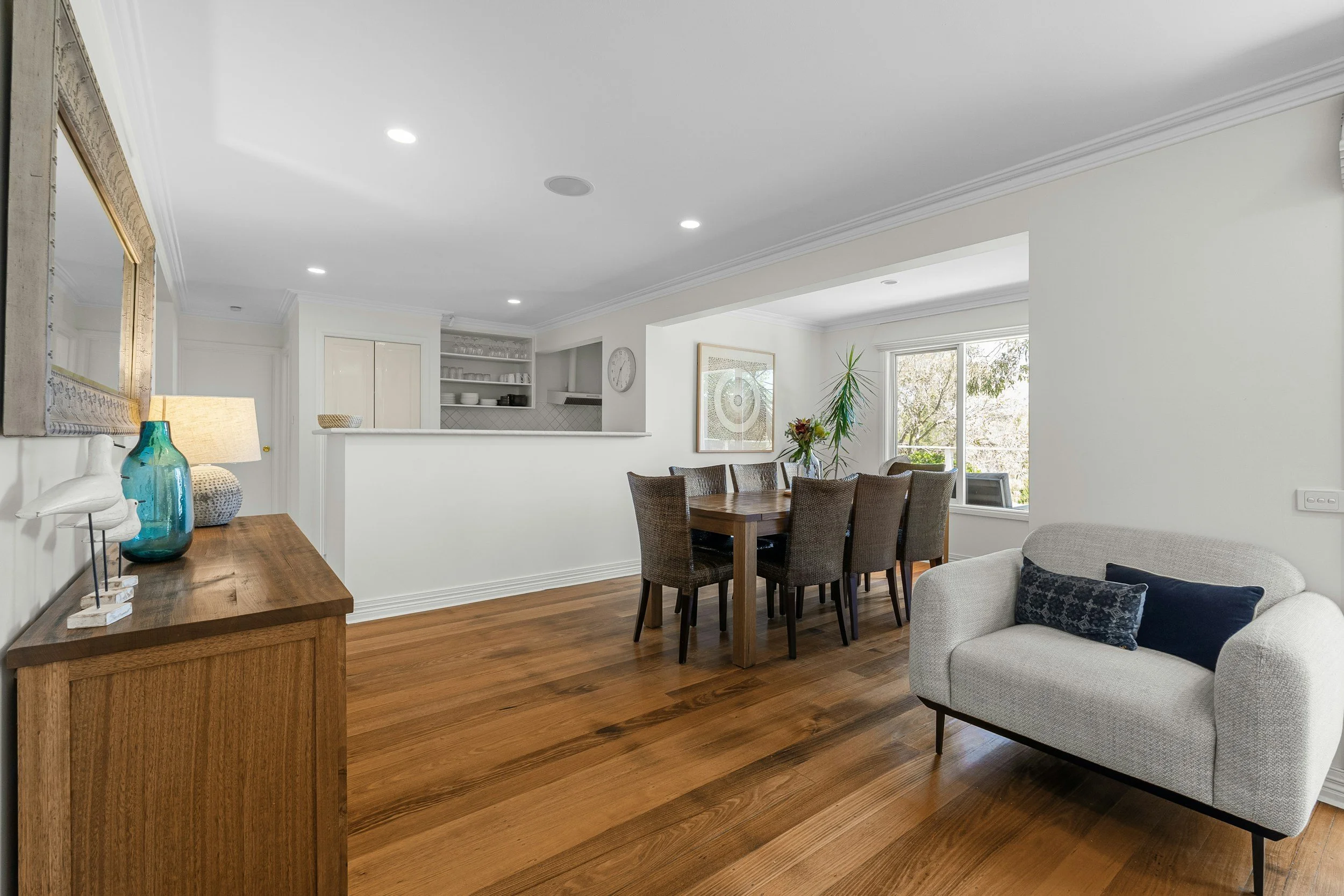Interior of a dining and living room area with hardwood floors, white walls, a grey armchair, an oval dining table with woven chairs, potted plants, a large window, and kitchen cabinets in the background.
