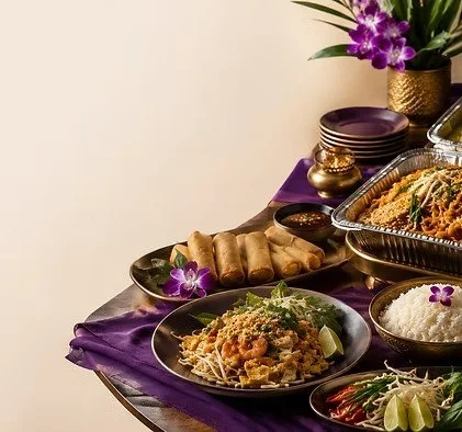 Assorted Asian dishes with spring rolls, rice, and noodle salad on a purple tablecloth with a flower arrangement in the background.