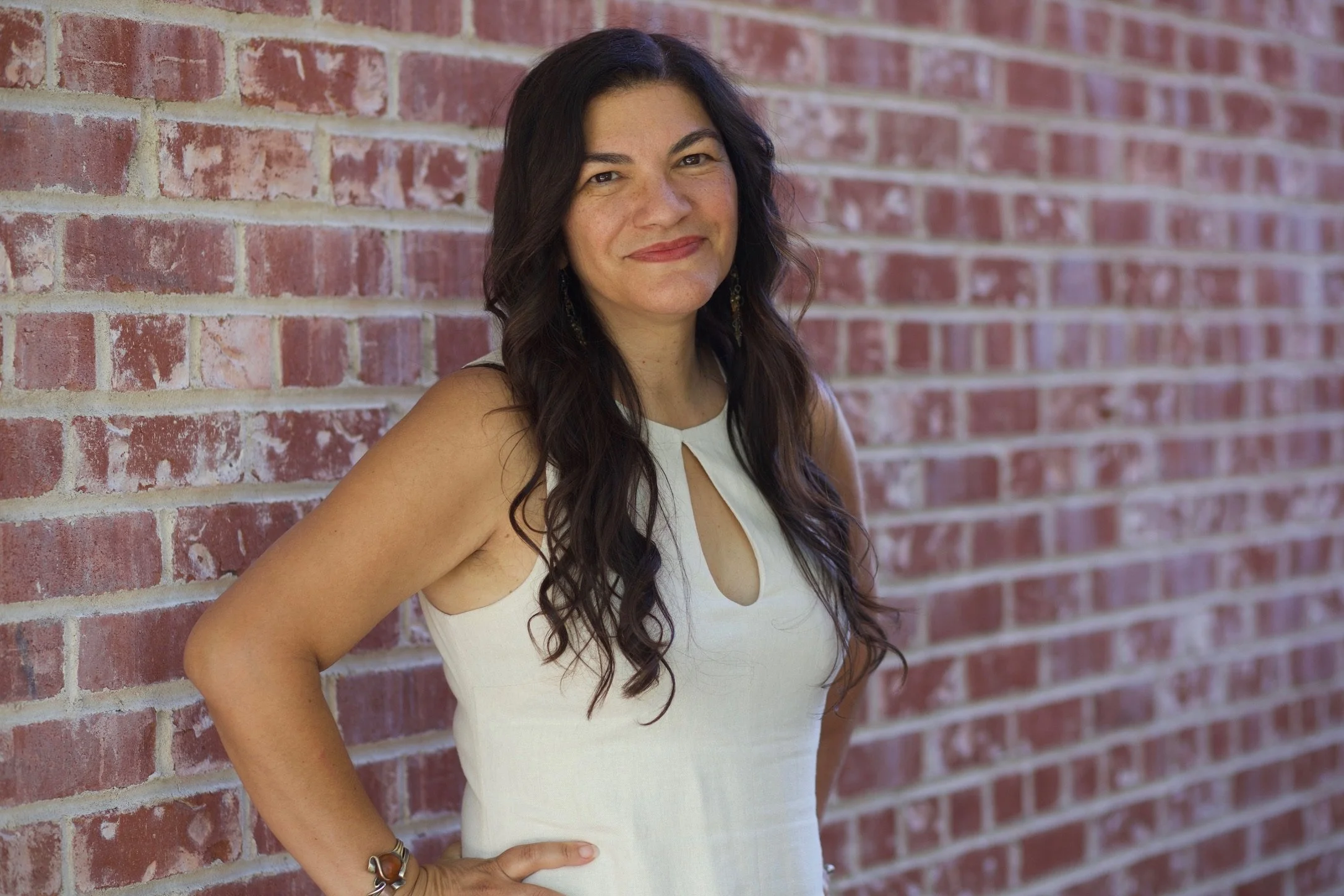 A woman with long dark wavy hair wearing a white sleeveless top, standing against a red brick wall, smiling at the camera with one hand on her hip.