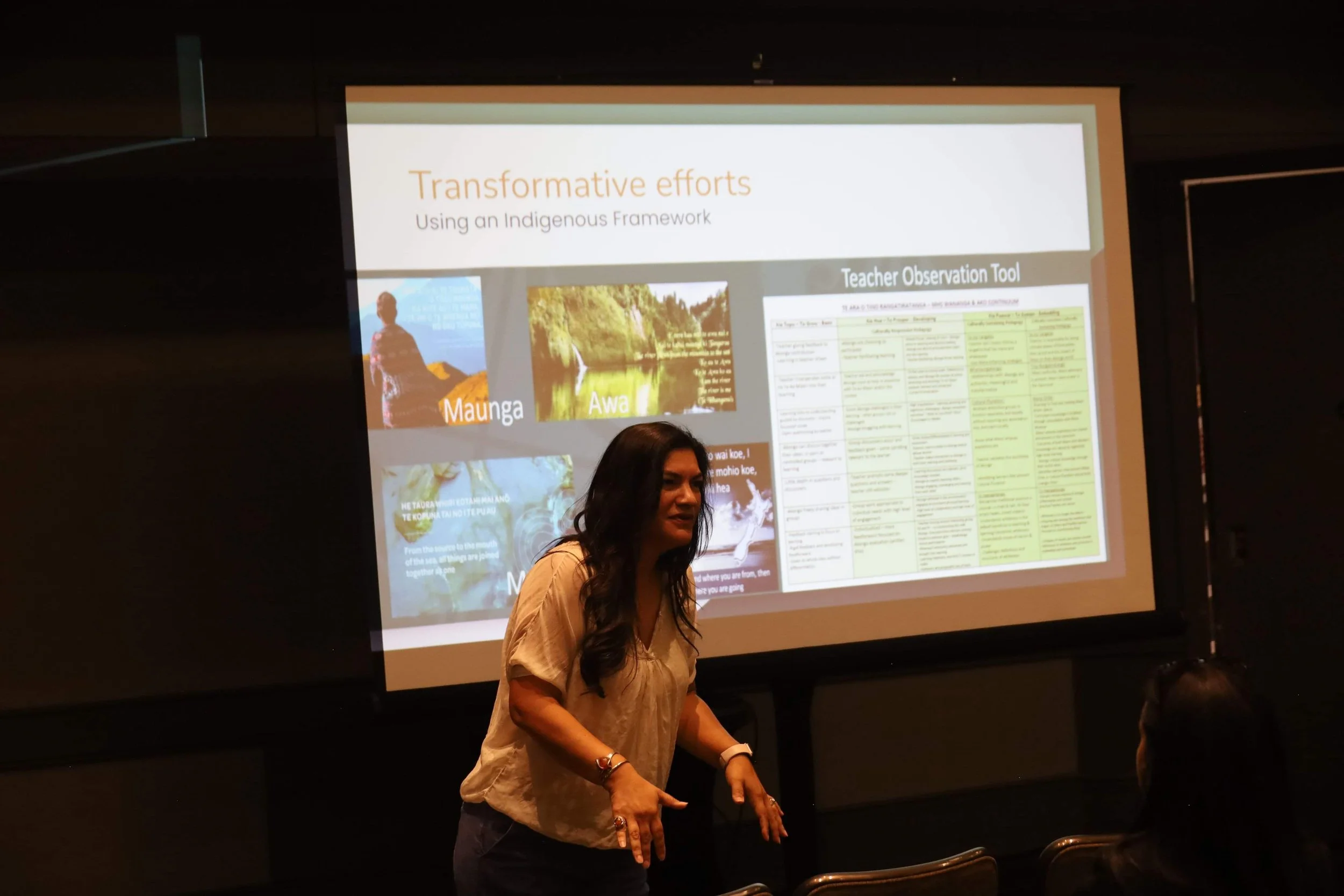 A woman giving a presentation in front of a large screen displaying slides about transformative efforts using an Indigenous Framework, including images of a person, nature scenes, and a teacher observation tool.