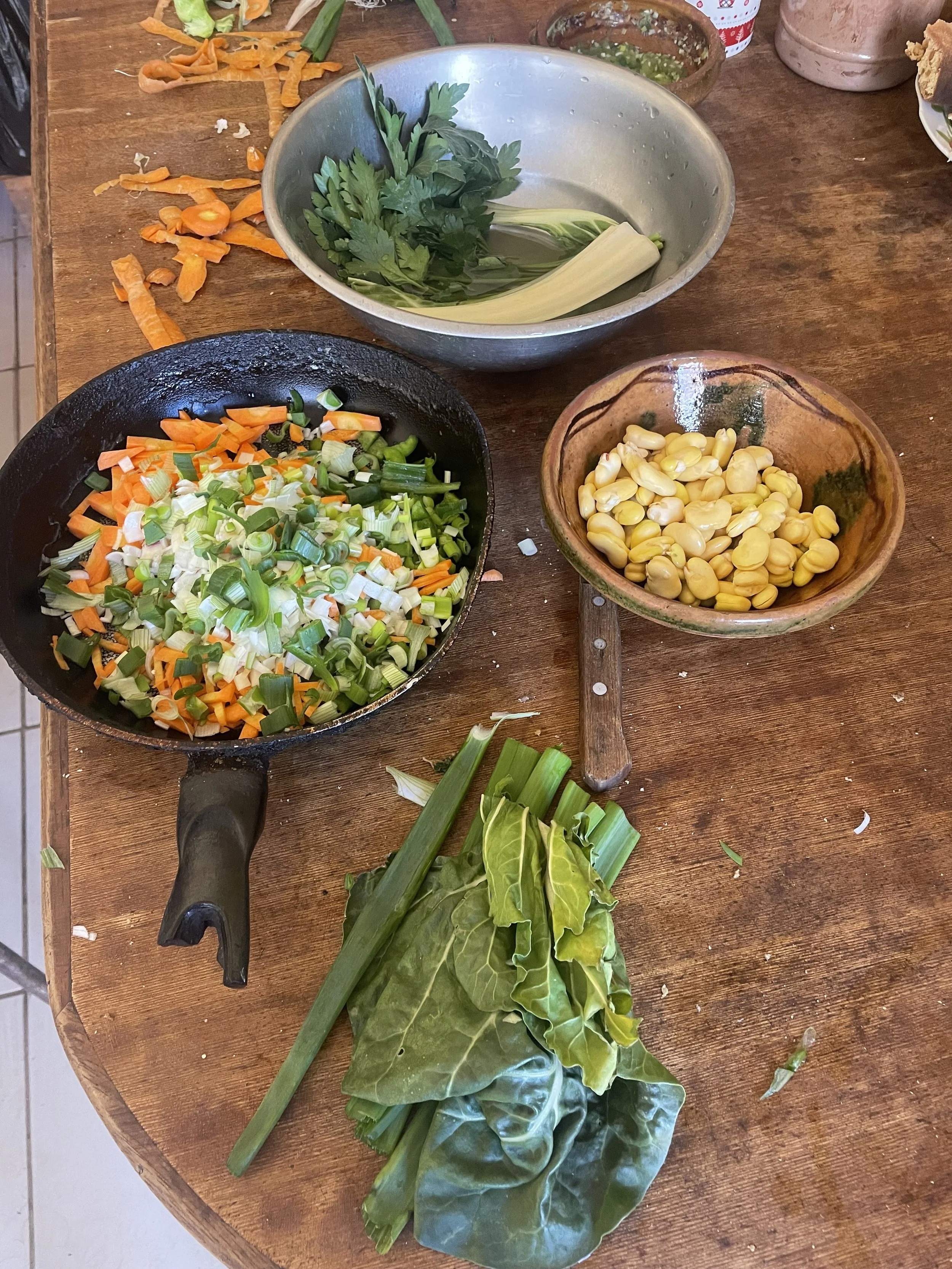 A wooden table with various chopped vegetables including carrots, green onions, cilantro, and leafy greens, along with a small bowl of corn kernels and a metal bowl with cilantro and lemon grass. There is also a black skillet, a wooden-handled spatula, and some scattered vegetable scraps.