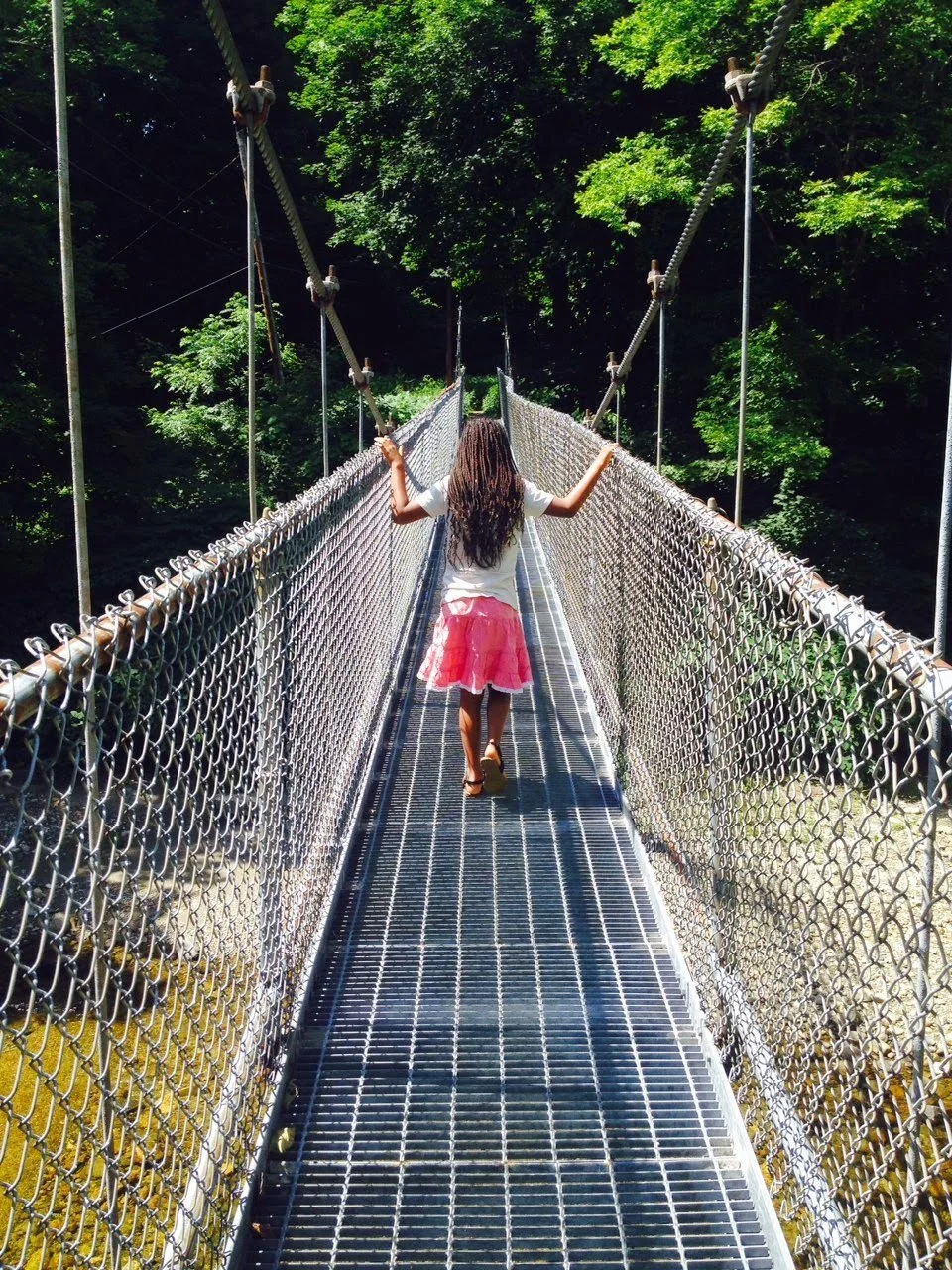 A girl with long curly hair, wearing a white shirt and pink skirt, walking on a narrow suspension bridge surrounded by lush green trees.