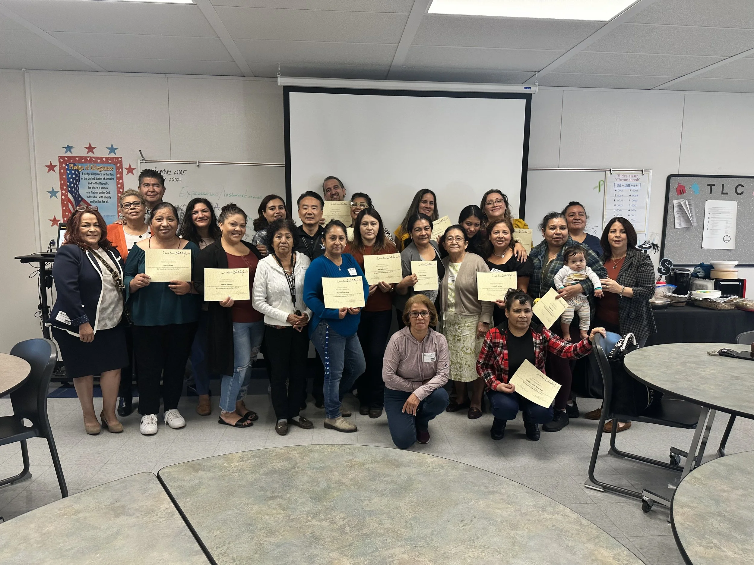A diverse group of people, mostly women, gathered together in a classroom, holding certificates and smiling for a photo. There are tables and chairs in the foreground, and a whiteboard and projector screen in the background.