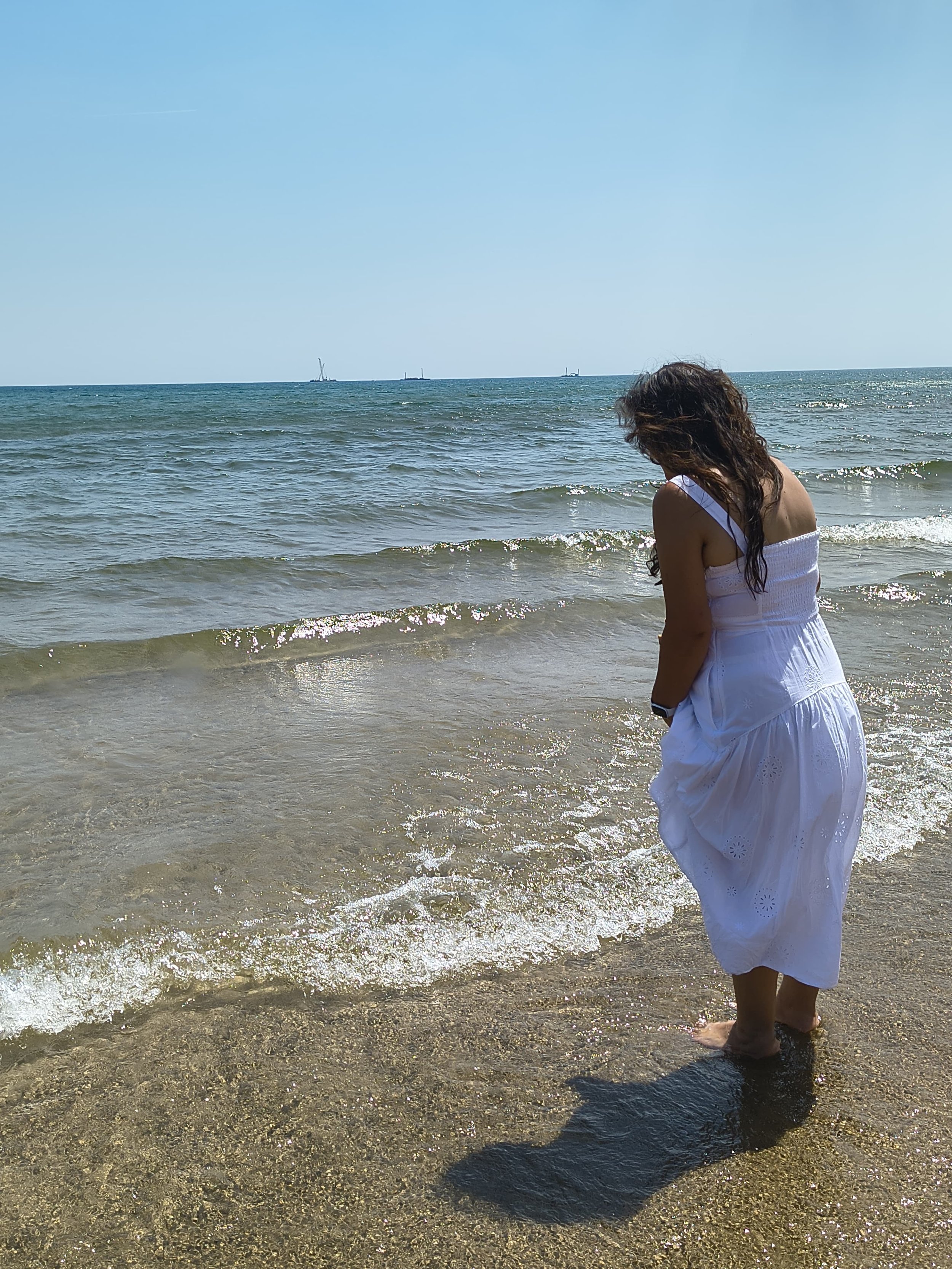 A woman with curly dark hair wearing a white dress stands at the edge of the water on a sandy beach, looking down at the ocean waves.