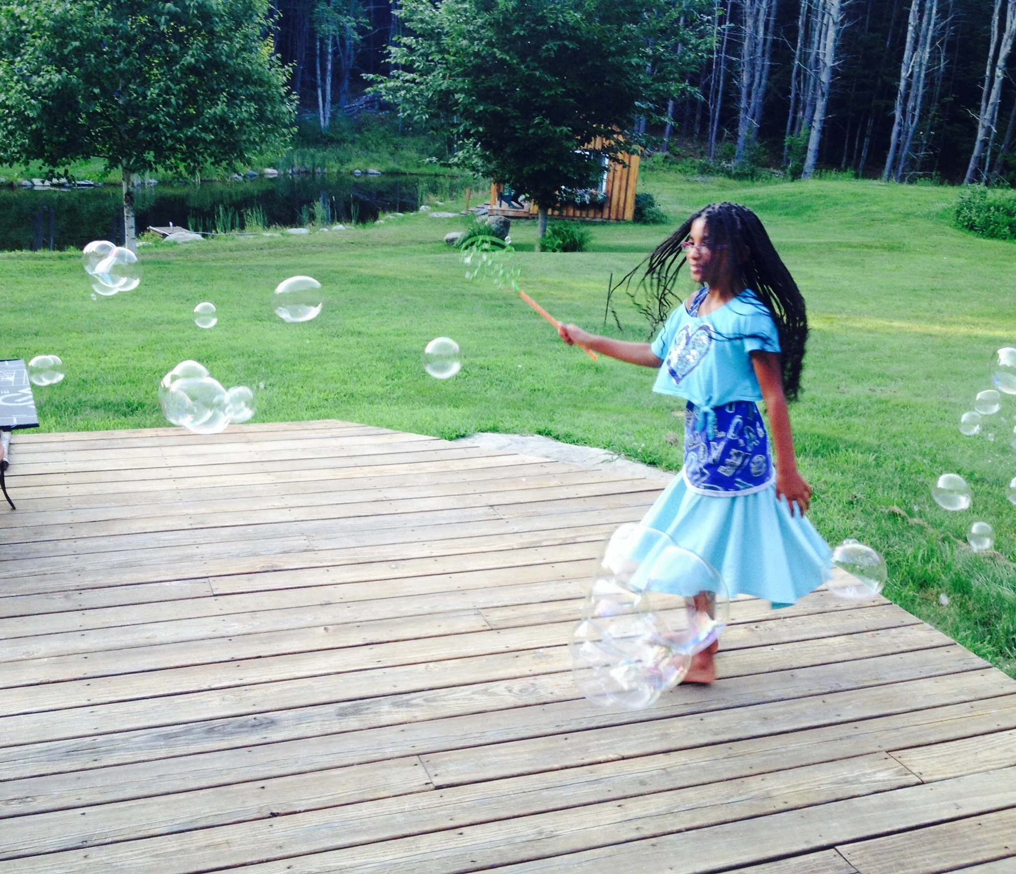 A young girl with long curly hair and glasses playing outside with bubbles on a wooden deck, surrounded by green grass, trees, and a small pond in the background.