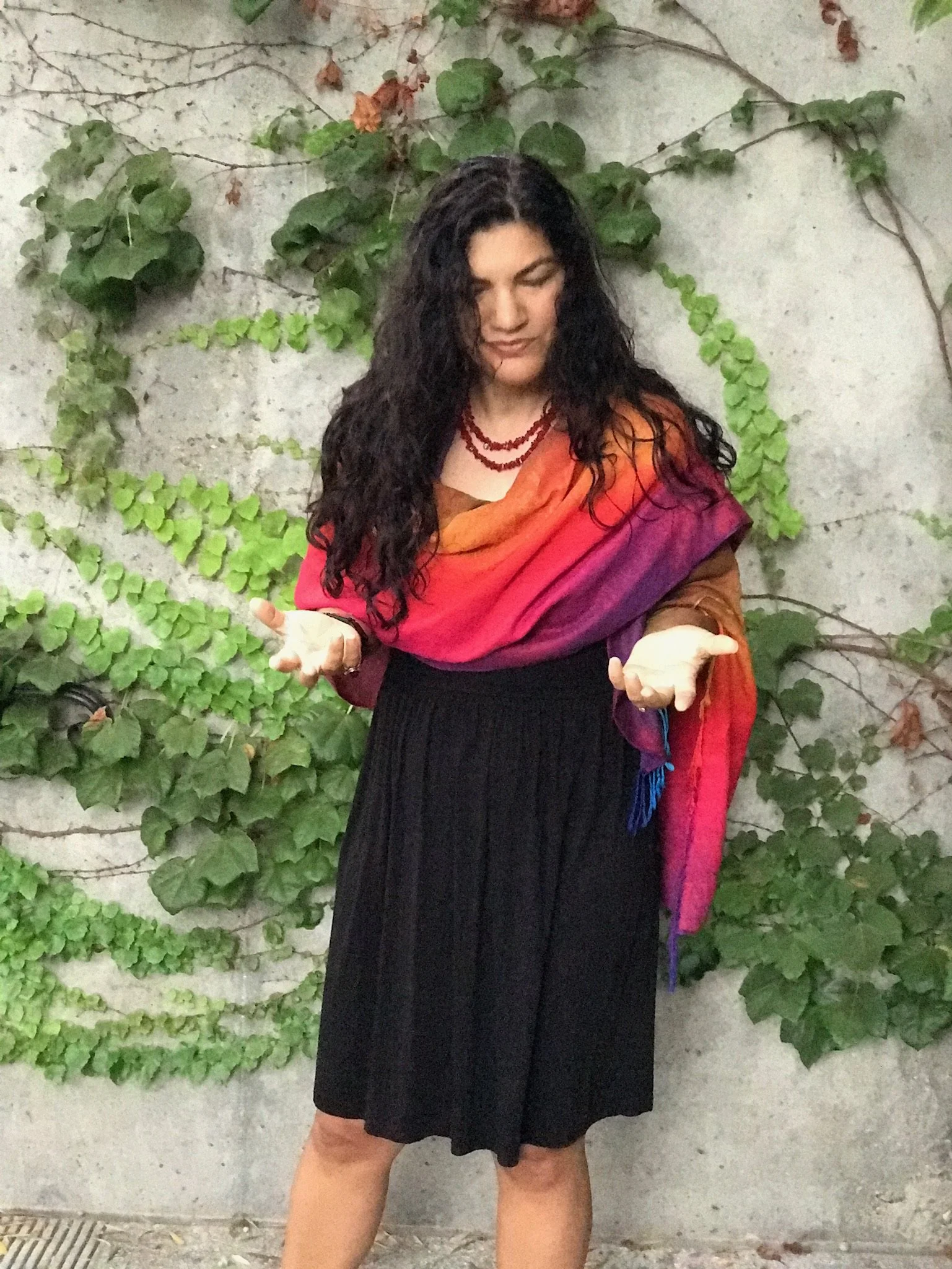 A woman with long curly dark hair is standing in front of a concrete wall covered with green ivy. She is wearing a colorful shawl, a black skirt, and multiple red necklaces. She has a neutral expression and is looking downward with her eyes closed, with her arms slightly extended and hands relaxed.