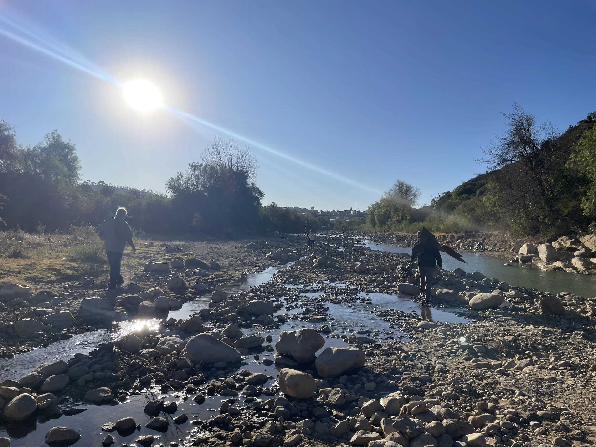 People hiking along a rocky stream with trees on either side under a bright sun in a clear blue sky.