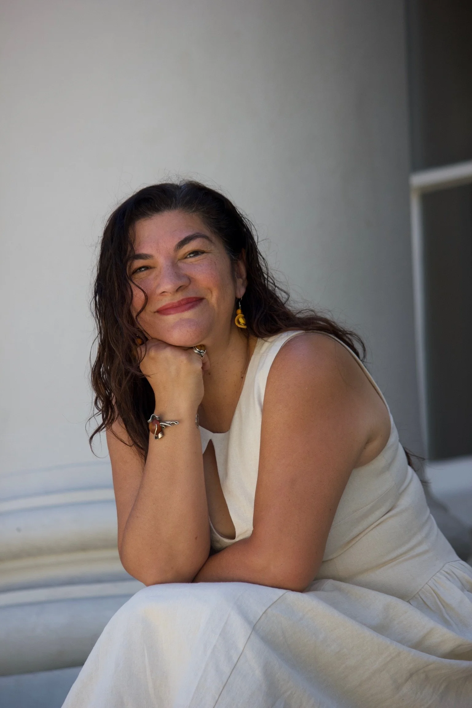 A woman with long, curly dark hair smiling and leaning her chin on her hand, sitting outdoors in a light-colored dress.