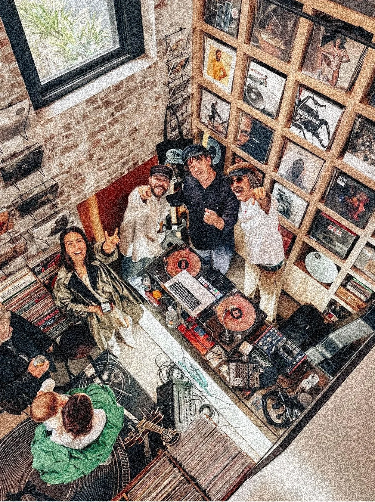 A group of people enjoying a DJ event in a cozy, brick-walled room decorated with framed pictures. The DJ is at the center with turntables and a laptop, surrounded by lively friends, some smiling and pointing at the camera.