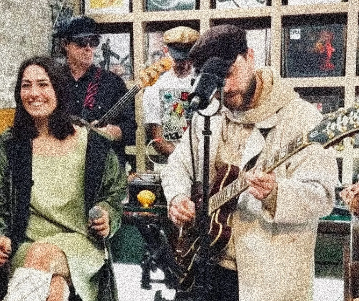 A group of four people, two men and two women, in an indoor setting with art on the walls. One woman is sitting, smiling, holding a microphone, while the man next to her is playing an acoustic guitar. The other woman is standing, wearing sunglasses and a hat, holding a bass guitar. The fourth person, in the background, is wearing a cap and holding a bass guitar.