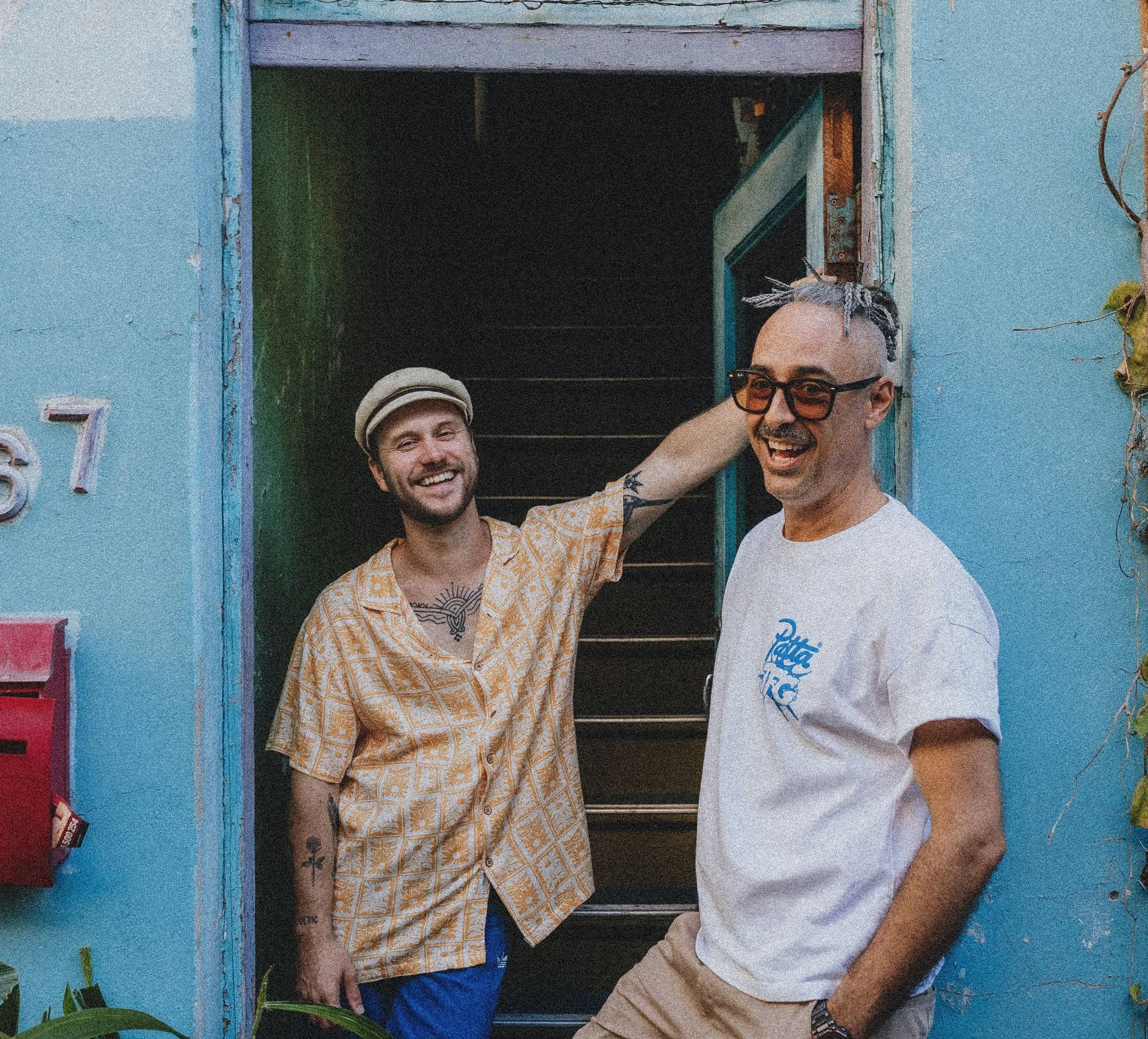 Two men smiling, standing in front of a weathered doorway with a dark interior. The man on the left wears a patterned yellow shirt, a beige cap, and has visible tattoos on his arm. The man on the right wears a white t-shirt with a graphic, sunglasses, and has short, styled hair with dreadlocks on top.
