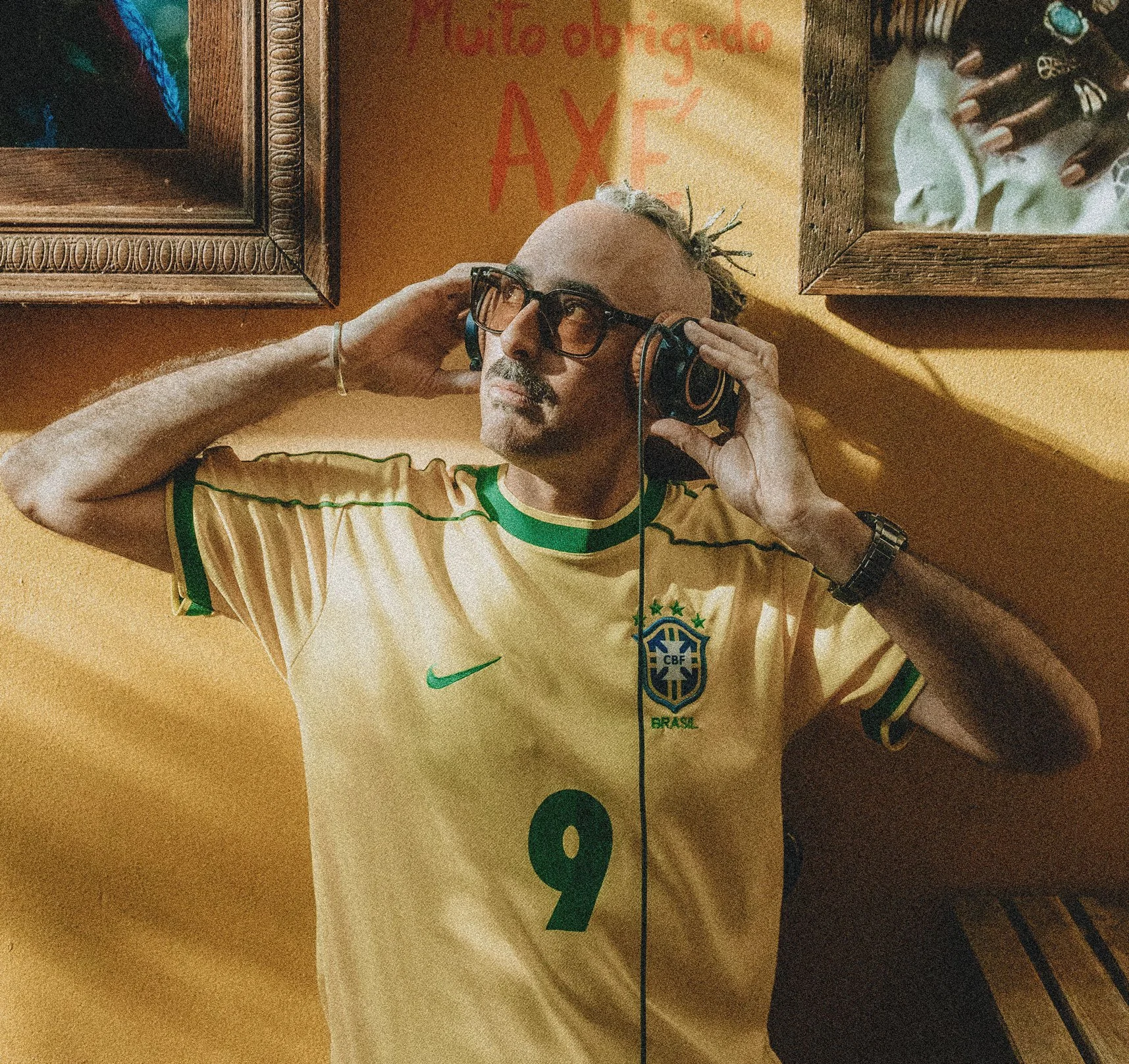 A person wearing glasses and a yellow Brazil national soccer team jersey lying on a wooden surface with headphones on, against a yellow wall with framed pictures and graffiti.