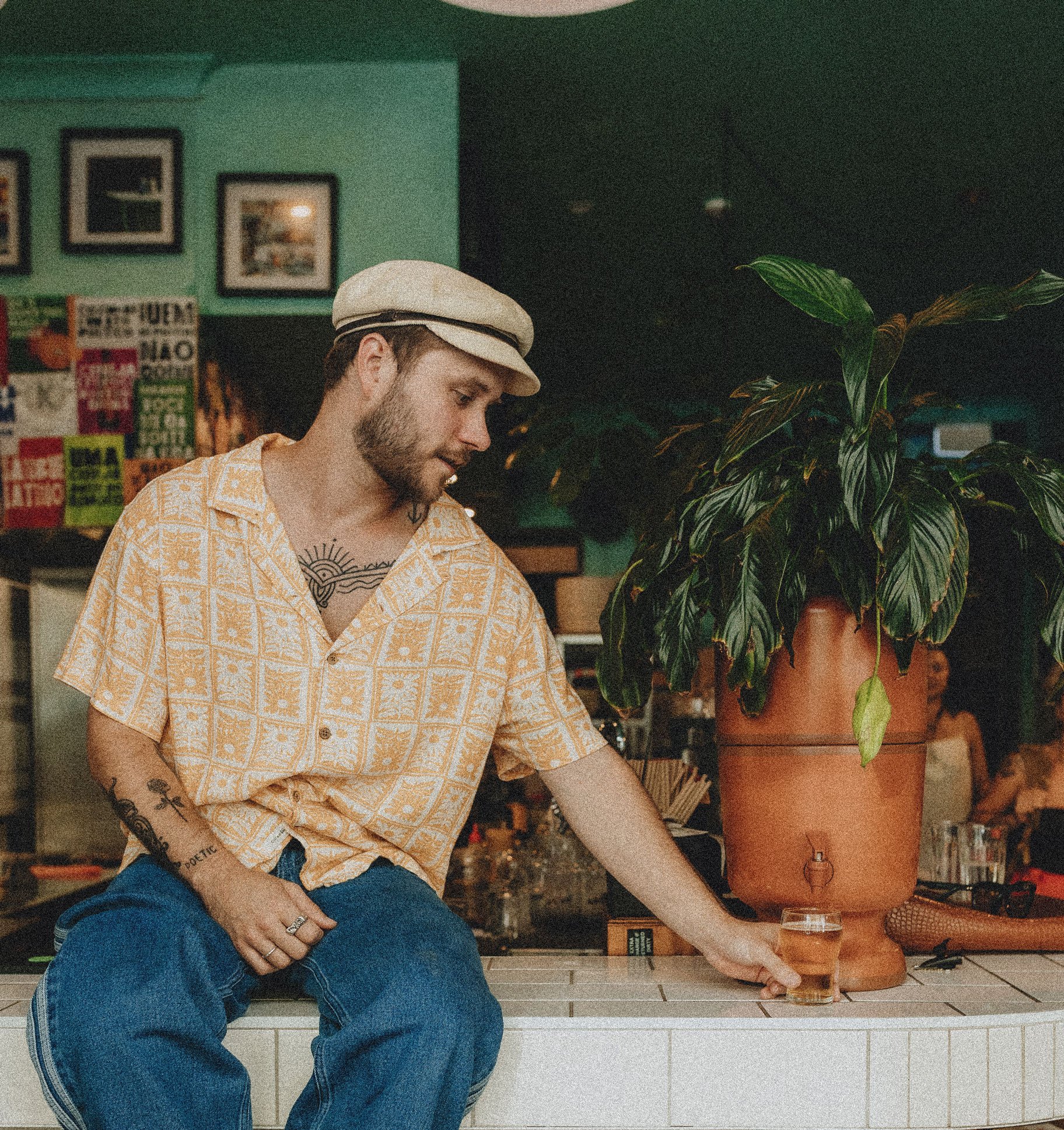 A man with a beard wearing a beige cap and patterned yellow shirt sitting on a counter, reaching for a glass of beer, next to a large potted plant in an indoor setting with framed pictures on the green wall.