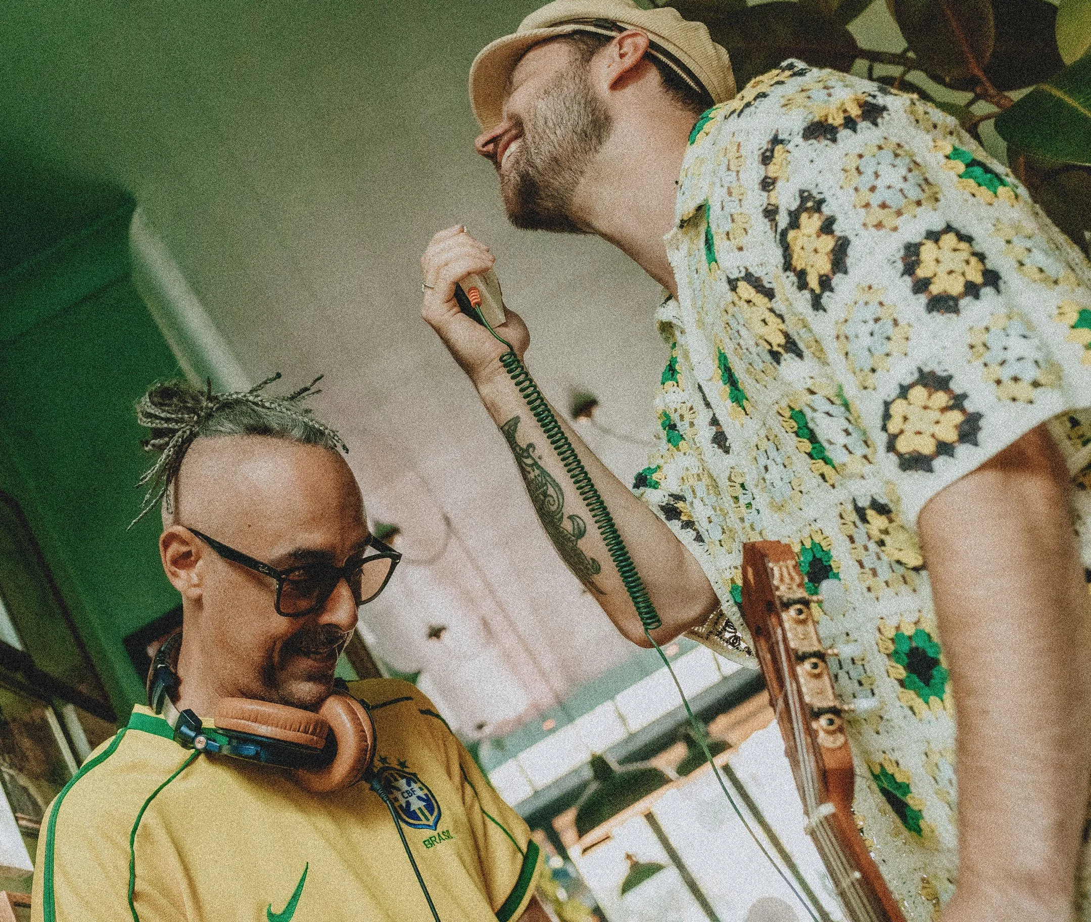 Two men in sports jerseys, one with a Brazilian flag patch, playing a game of foosball. One man is wearing glasses and headphones, the other is smiling, wearing a cap and a colorful shirt.