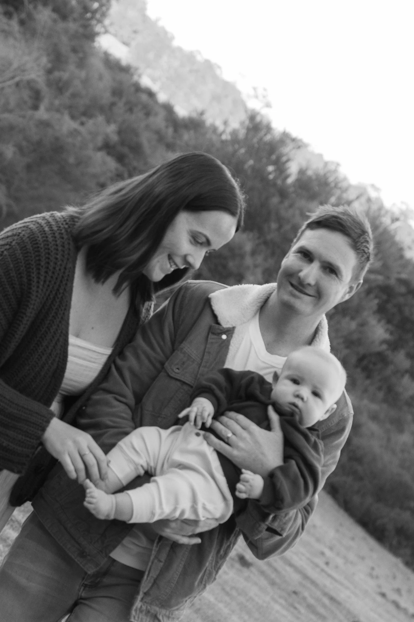 A black and white photo of a smiling woman, a young man, and a baby outdoors with trees in the background. The woman is looking down, the man is looking at the camera, and the baby is looking forward.