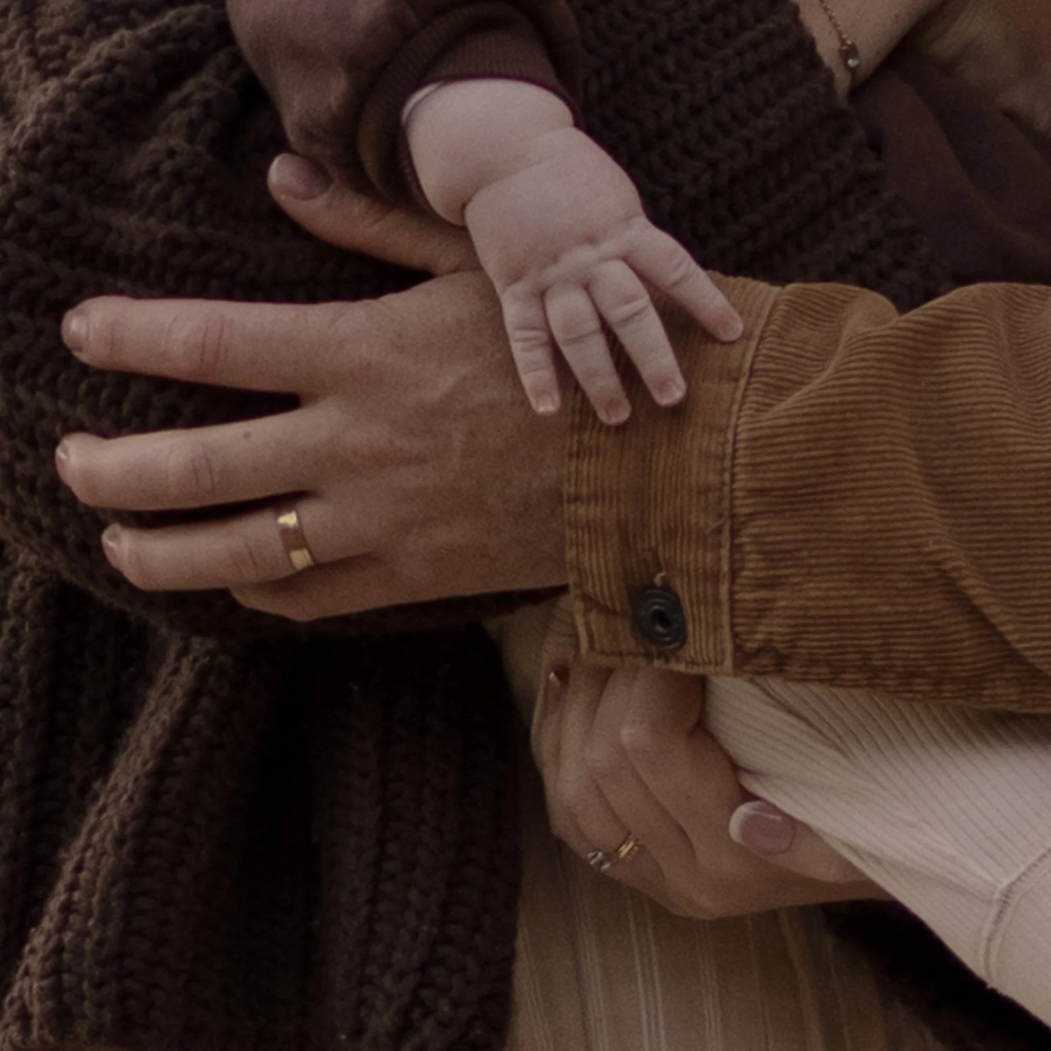 Close-up of hands with one hand holding a baby finger, showing jewelry including rings and a bracelet. The background includes clothing textures and a wooden surface.