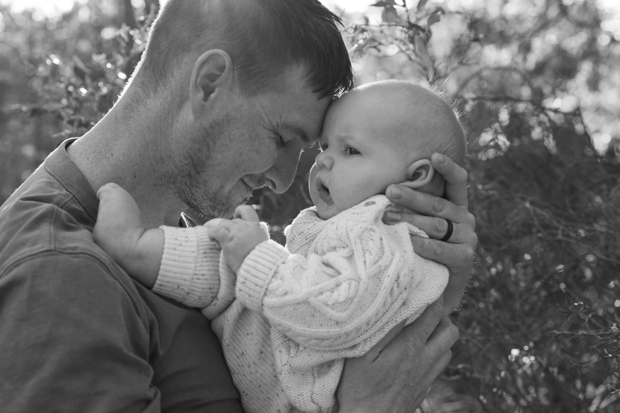 A black and white photo of a man holding a baby close, their foreheads touching, outdoors with trees in the background.