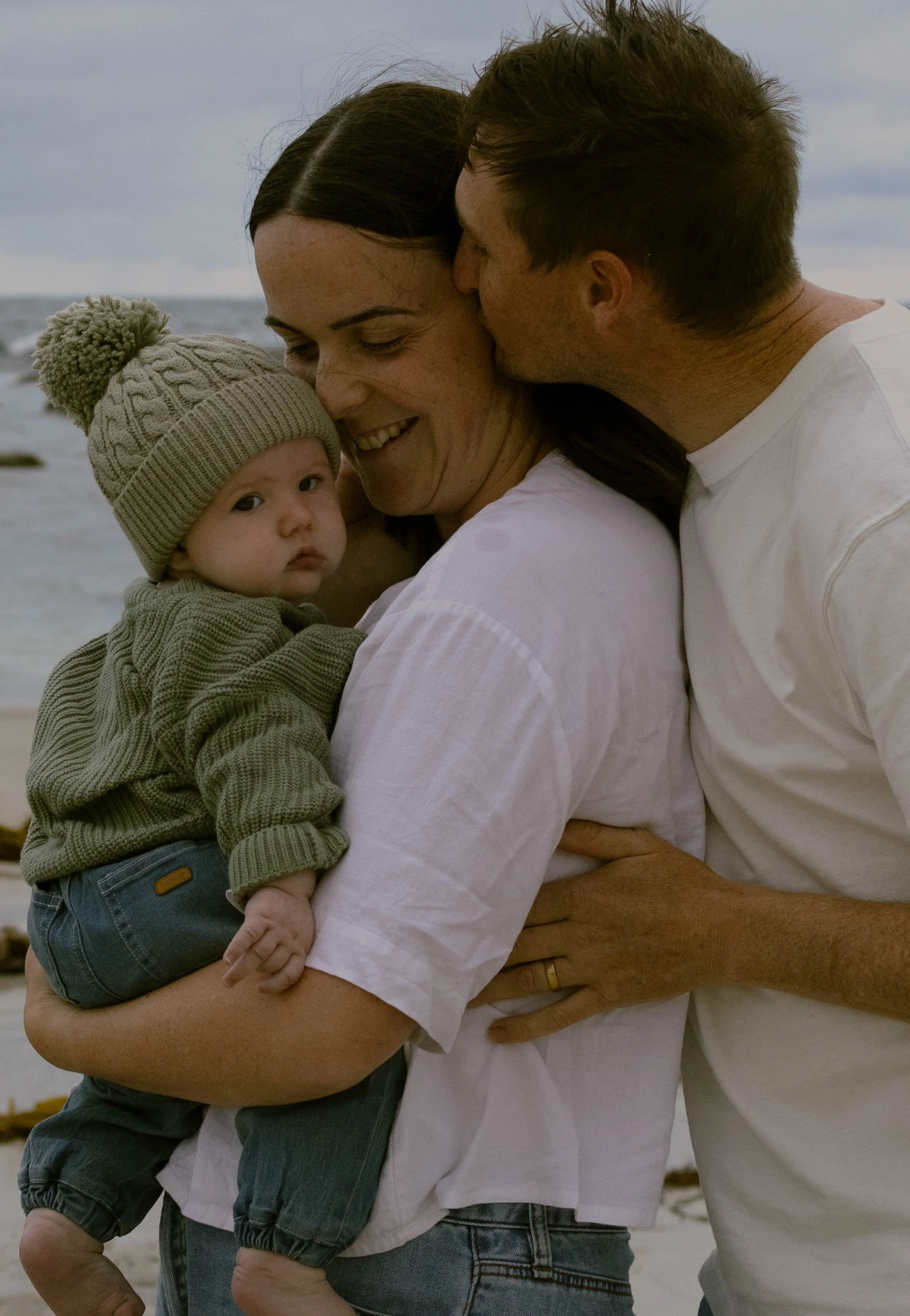 A family of three at the beach, with a woman holding a young child and a man kissing her on the cheek. The woman has brown hair, and the child is wearing a beige knit hat. The man has messy brown hair. All are dressed casually in white and gray cloth