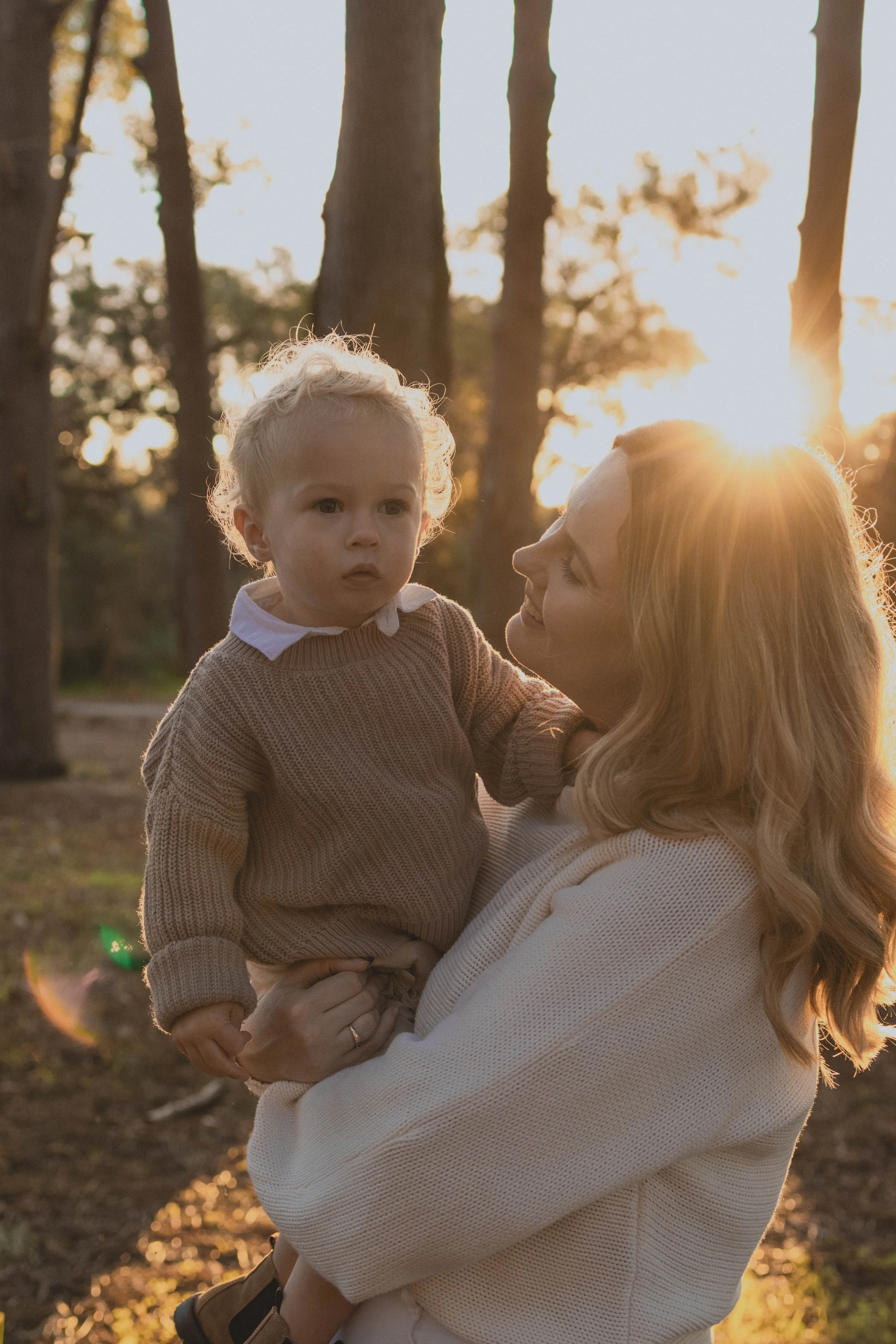 A woman holding a young child outdoors during sunset or sunrise with tall trees in the background.