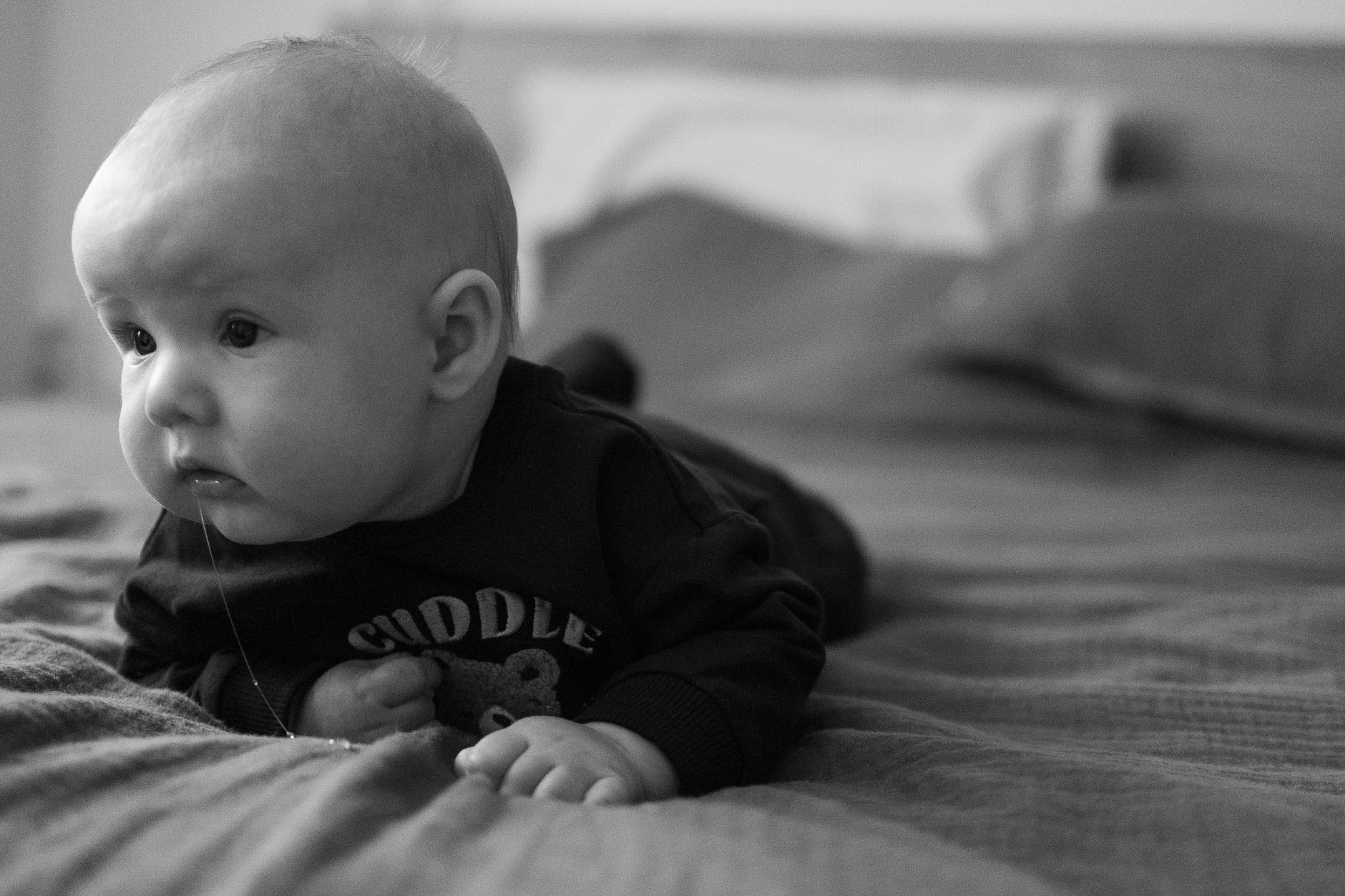 A young child with a shaved head lying on their stomach on a soft surface, looking intently to the side, with a blurred background.