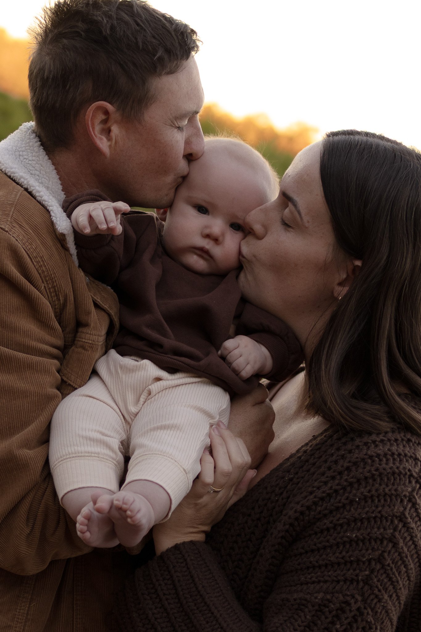 A family of three sharing a tender moment outdoors at sunset. The father kisses the baby's head, and the mother kisses the baby's cheek. The baby, with blonde hair, looks at the camera.