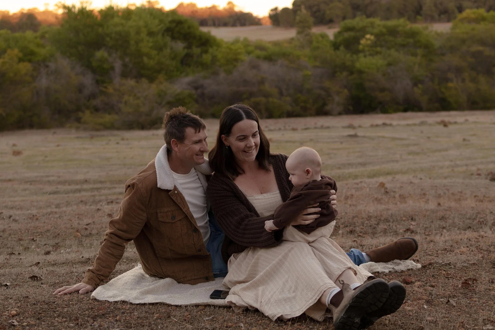 A family of three, a man, woman, and baby, sitting on the ground outdoors during sunset, smiling and enjoying each other's company.
